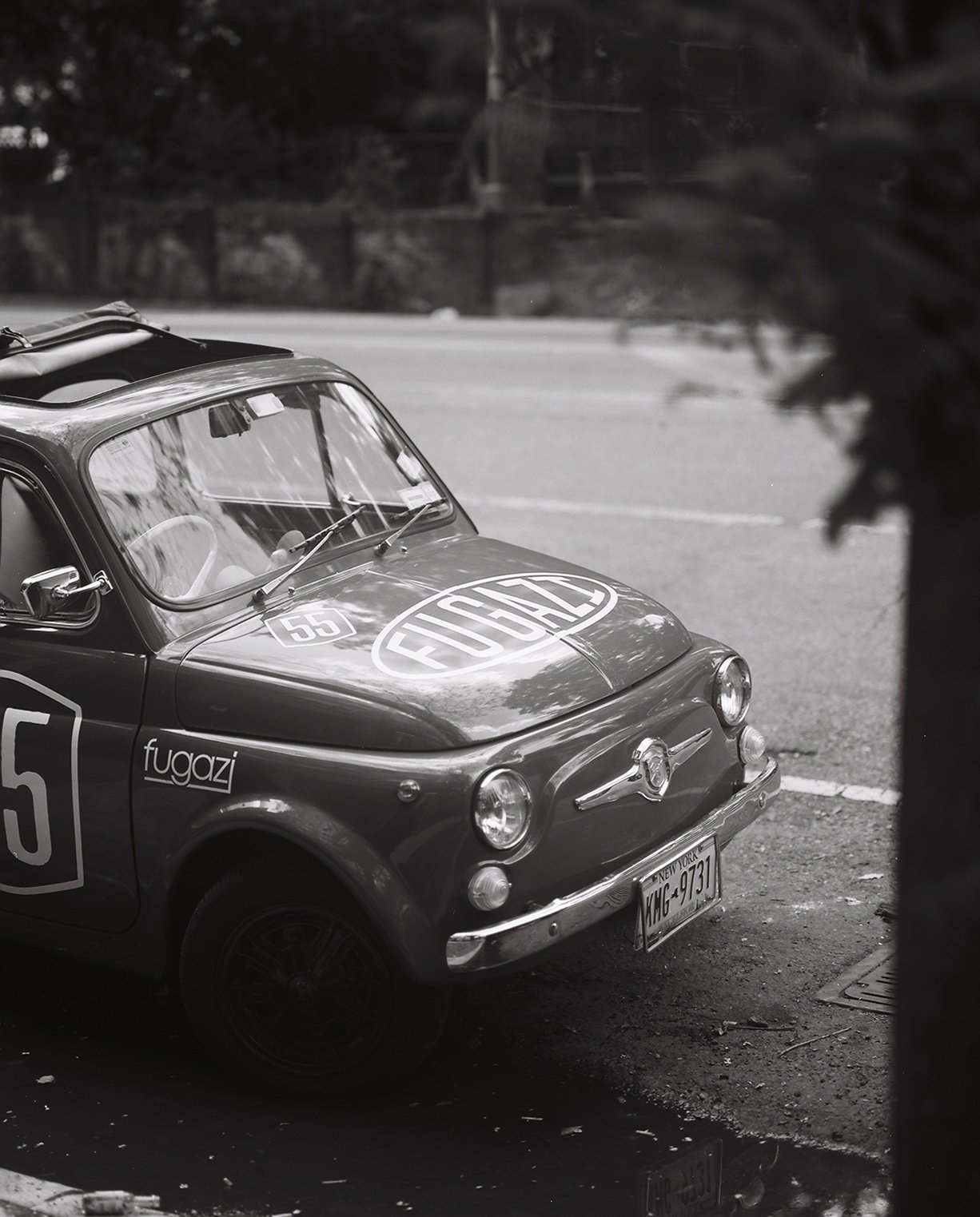 A vintage racing car with a large 'FUGAZI' logo on the hood and a number '55'. It has a classic, rounded design, and is parked on the side of a road. The car is identified as a Fiat, with a New York license plate reading 'MHG 9731'. The surrounding s
