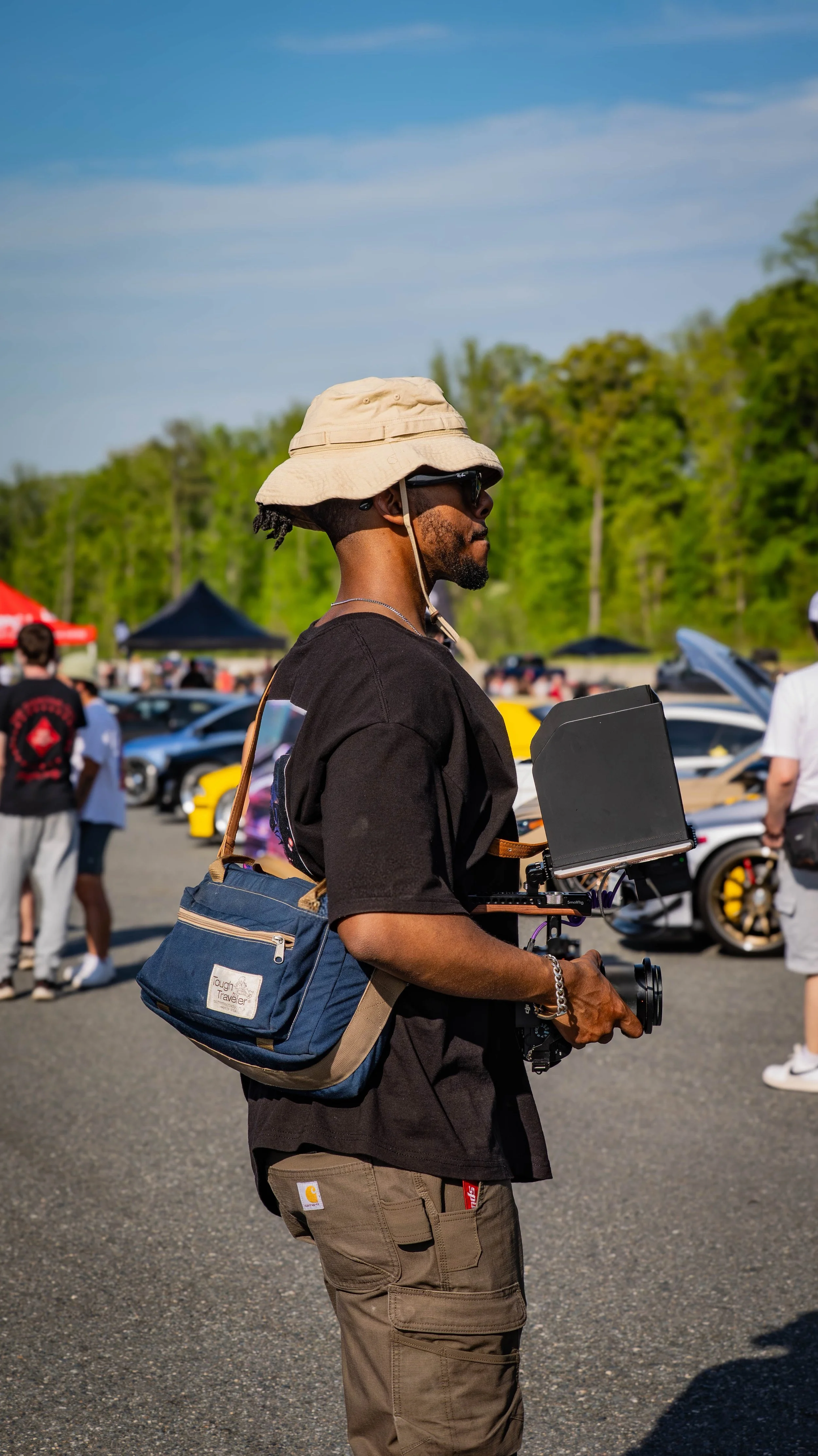 A man wearing a beige bucket hat, sunglasses, a black T-shirt, and olive green cargo pants, is holding a camera with a stabilizer in an outdoor car show with various cars and people in the background.