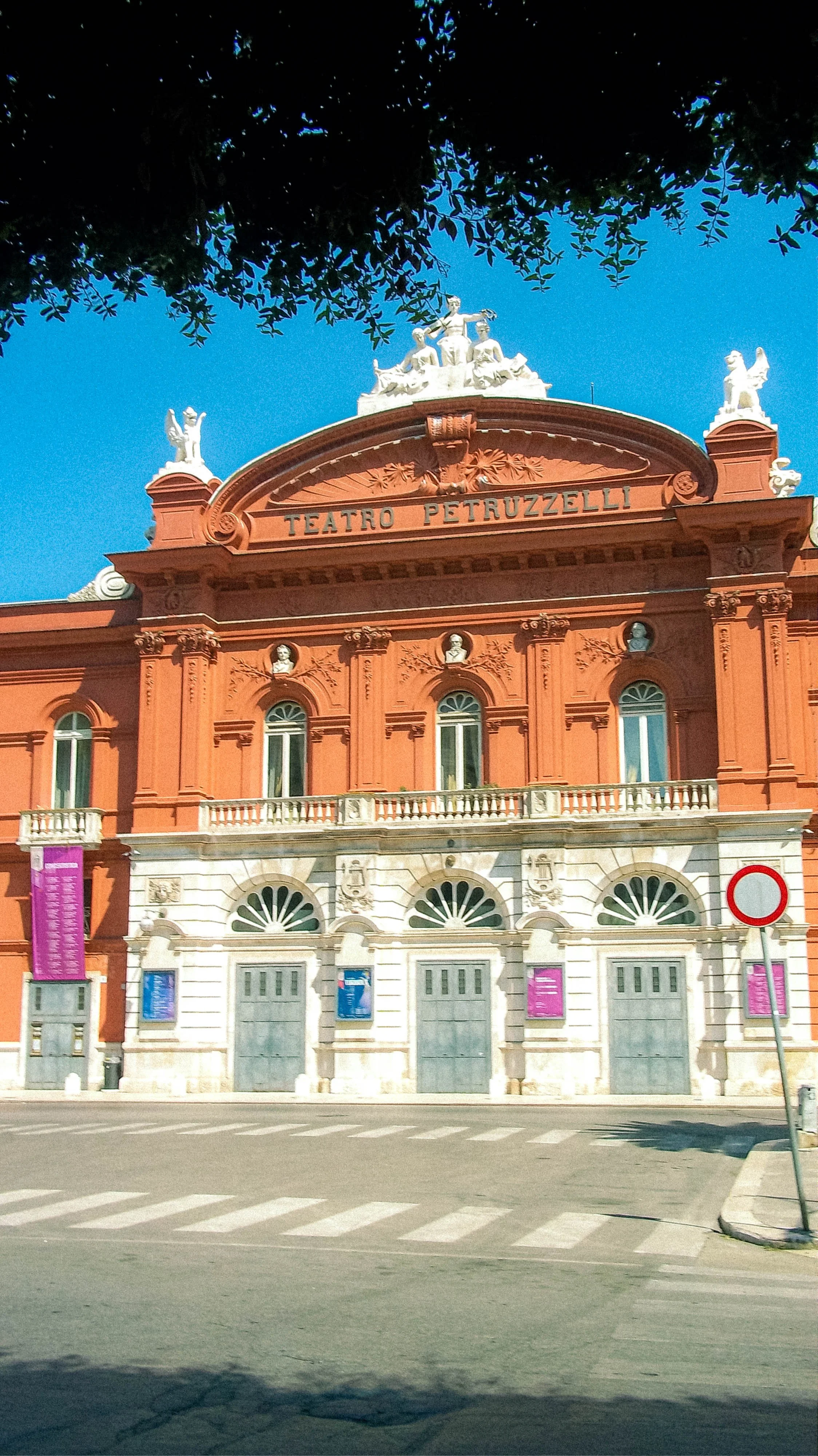 The building has an orange facade with classical architectural details, including decorative columns and sculptures on the roof. It has a sign that reads 'Teatro Petruzzelli' and colorful banners hanging from the lower windows. There are trees at the