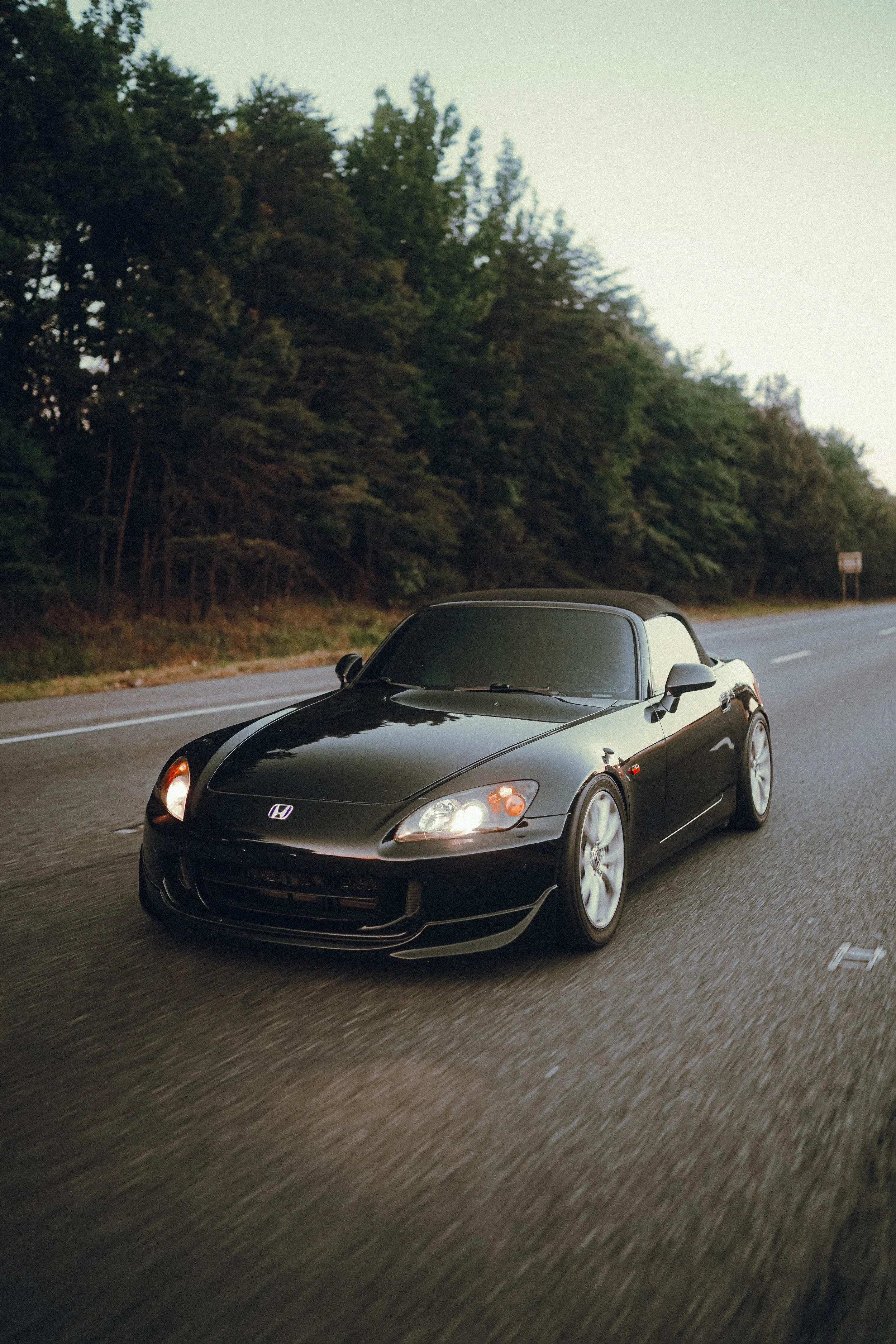 A black Honda S2000 sports car driving on a highway with trees on the side during sunset.