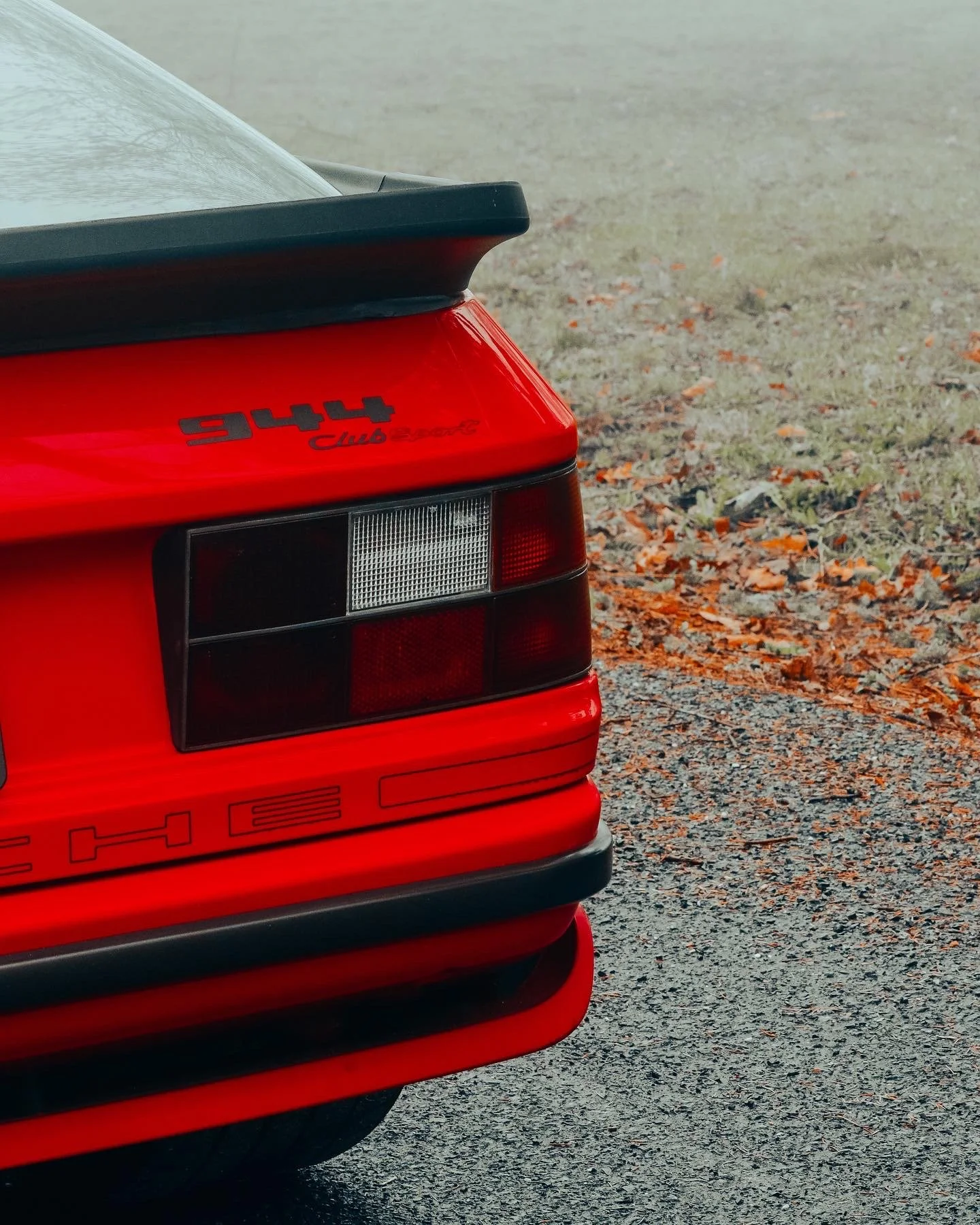 Rear close-up of a red Porsche 944 sports car parked on a gravel surface with fallen leaves, showing the car's tail light, rear spoiler, and model badge.