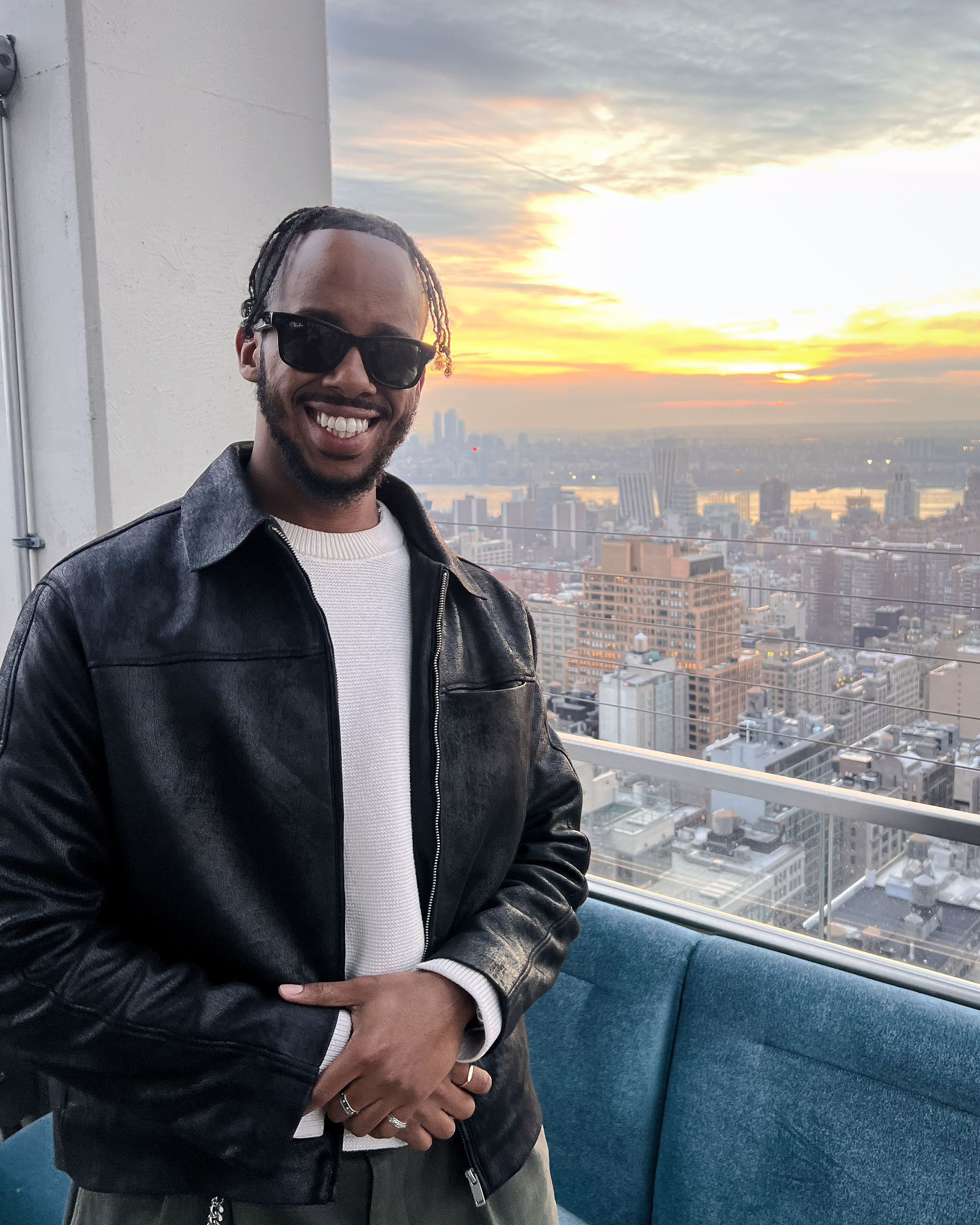 A man wearing black sunglasses and a leather jacket smiling, standing in front of a city skyline at sunset, with tall buildings and a river visible in the background.