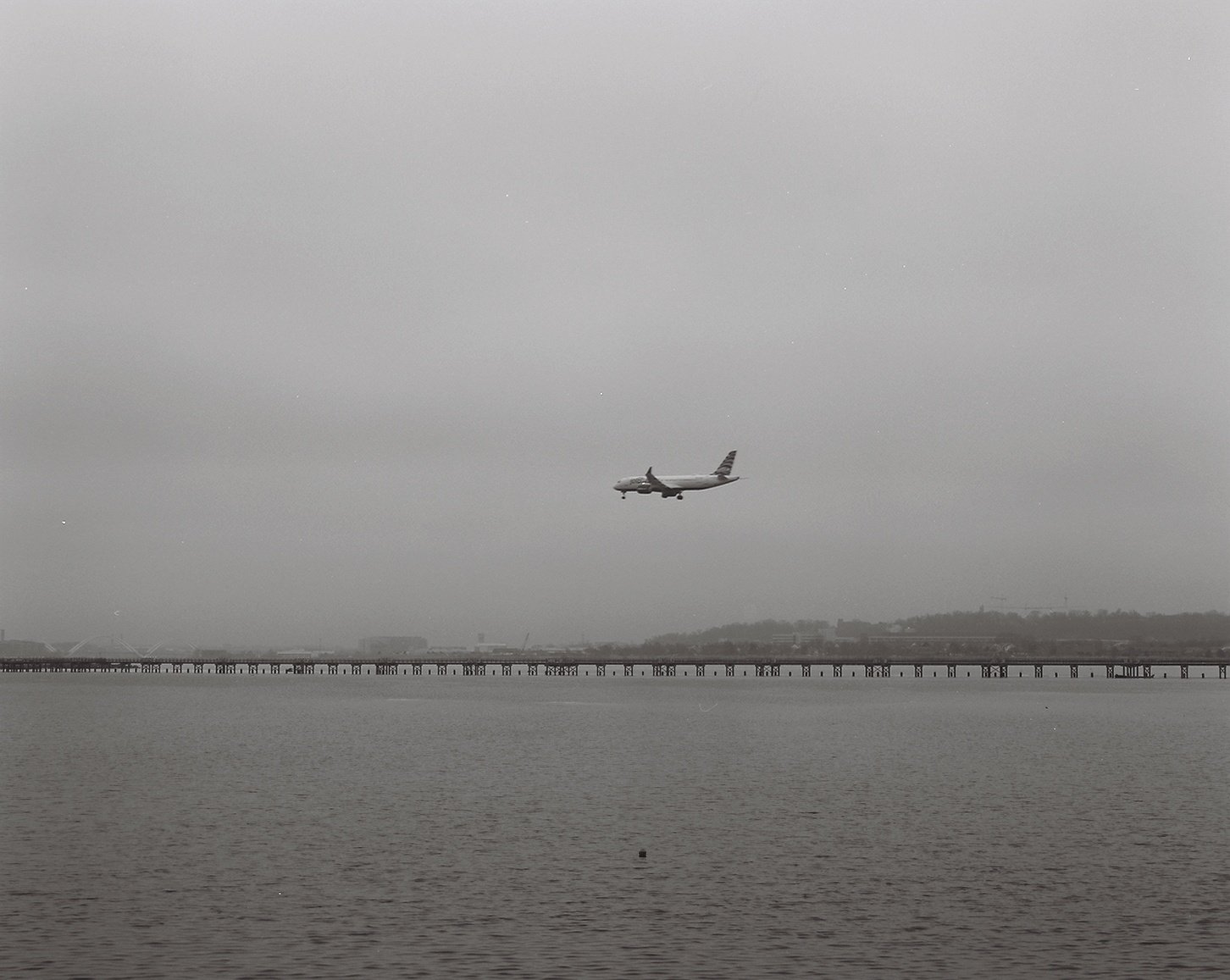 Gray sky with a commercial airplane flying above a body of water, with a distant shoreline and a bridge or pier extending across the water.