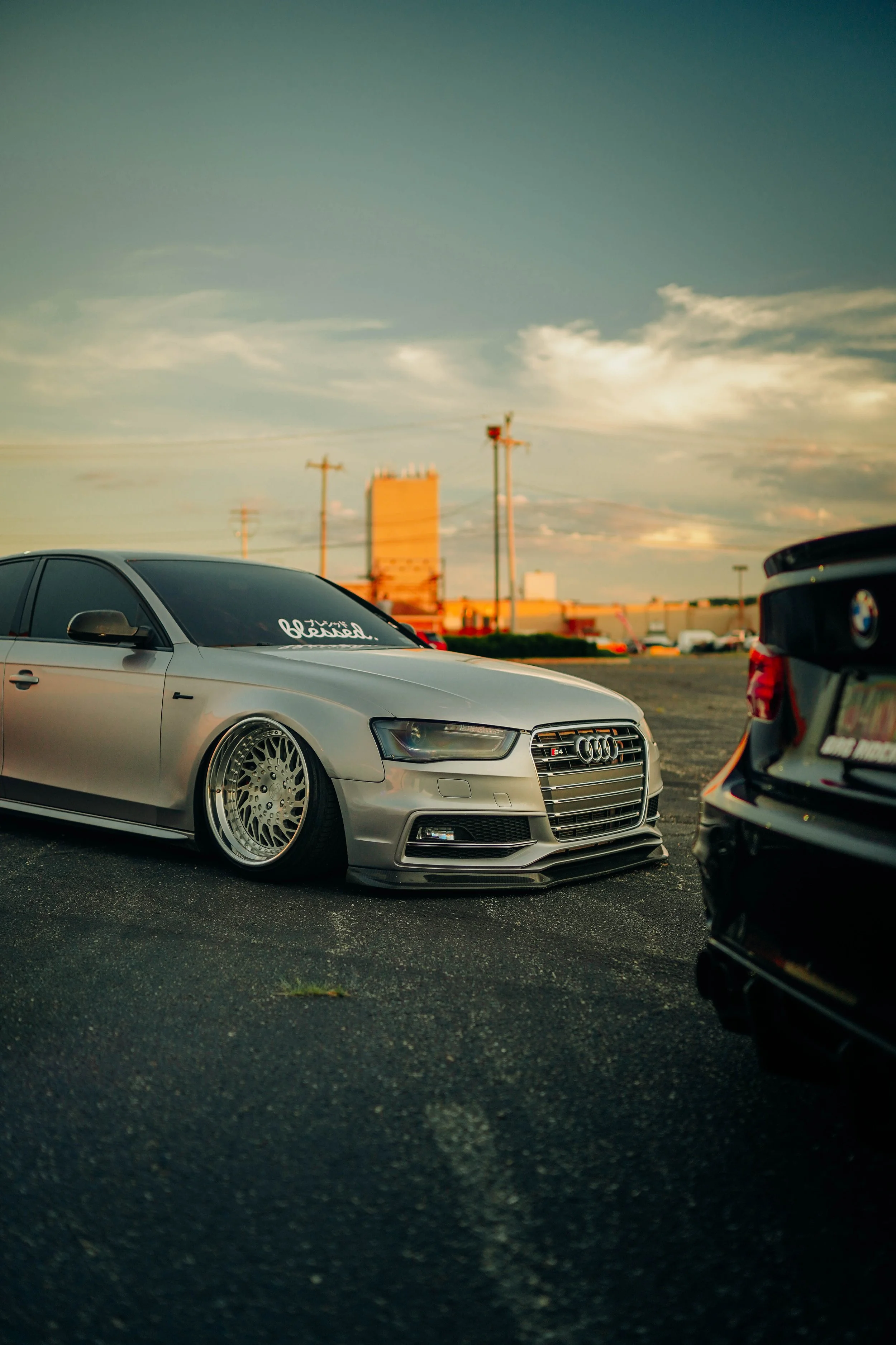 A silver Audi sedan with custom wheels parked at a car meet during sunset, with other cars and a parking lot visible in the background.