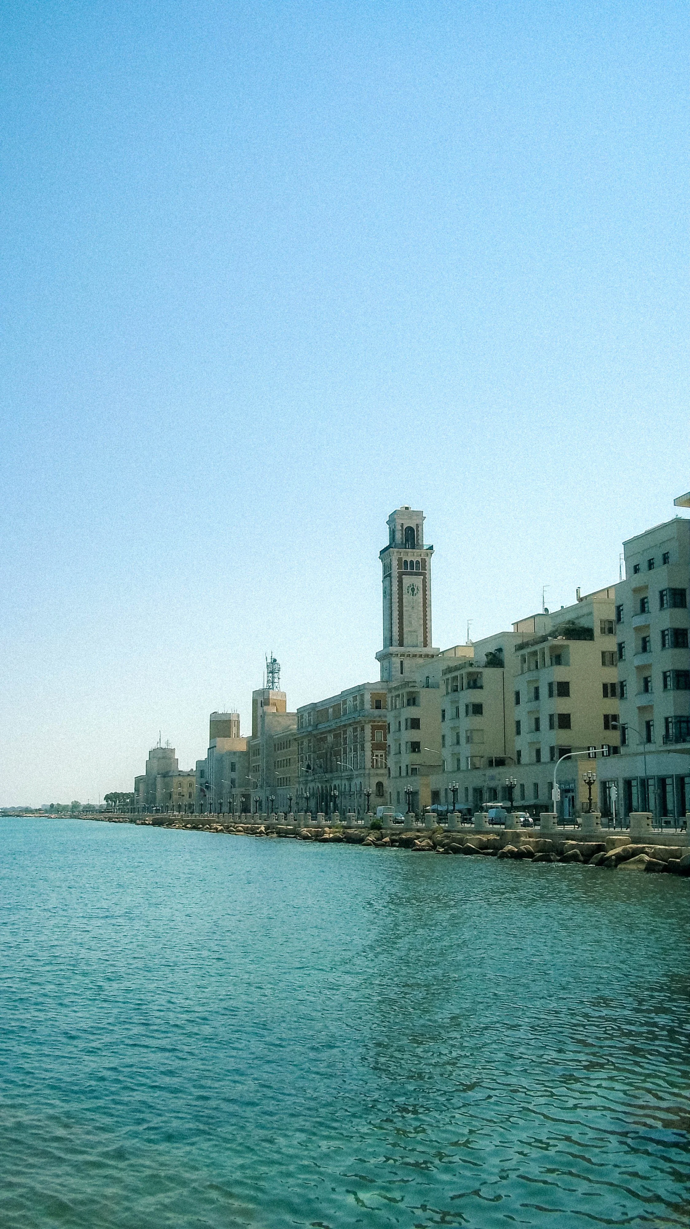 Cityscape along a waterfront with historic buildings and a tall clock tower under a clear blue sky.