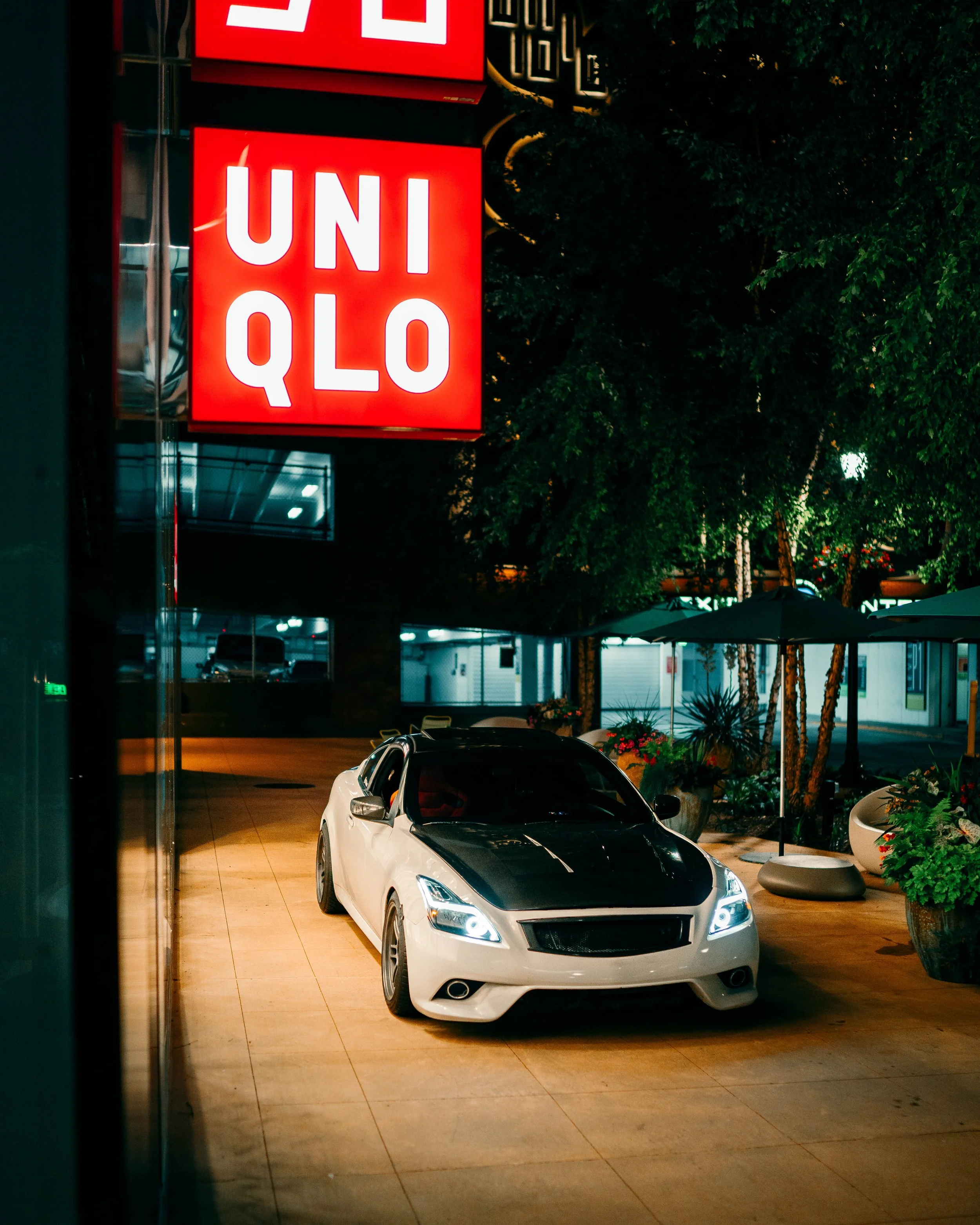 A white and black sports car parked inside a building or showroom, with a red illuminated UNIQLO sign above it. The scene is nighttime, with trees and outdoor seating visible outside the building.