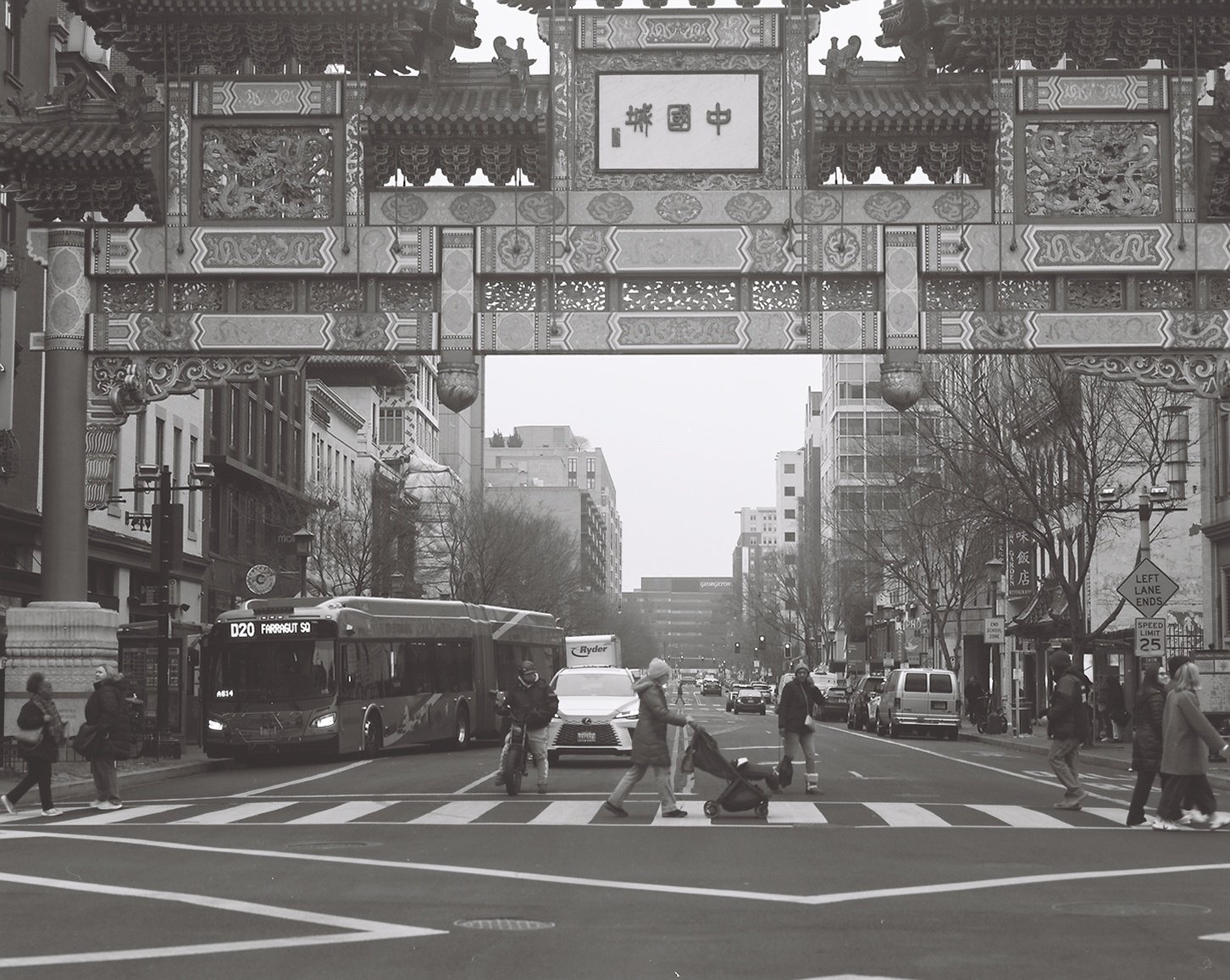 A busy city street scene with pedestrians crossing the crosswalk, a bus, cars, and buildings, under a decorative Chinese-style archway in black and white.