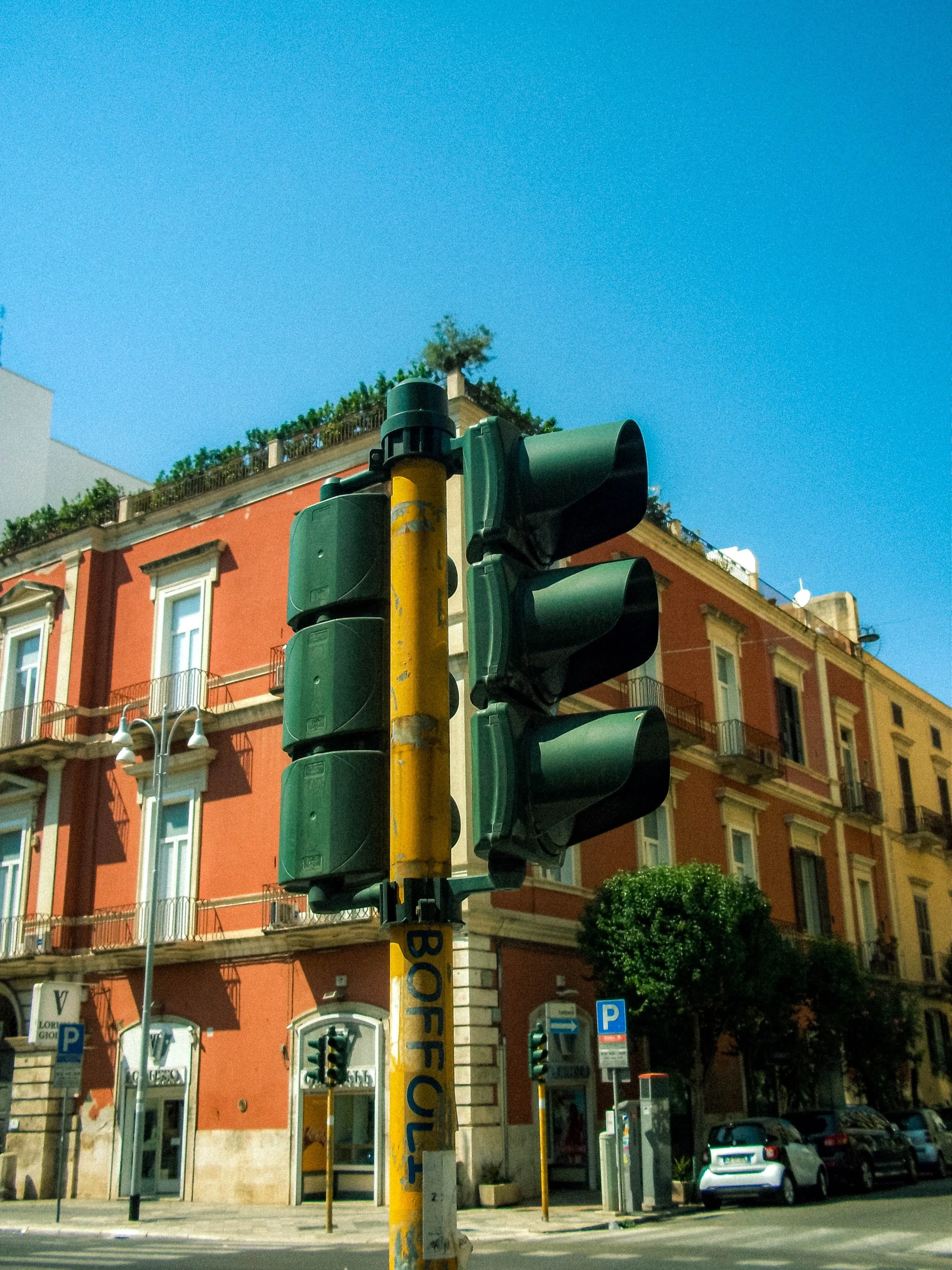 Traffic light with multiple signals in front of a colorful building with balconies, windows, and greenery on the rooftop, and parked cars on the street.