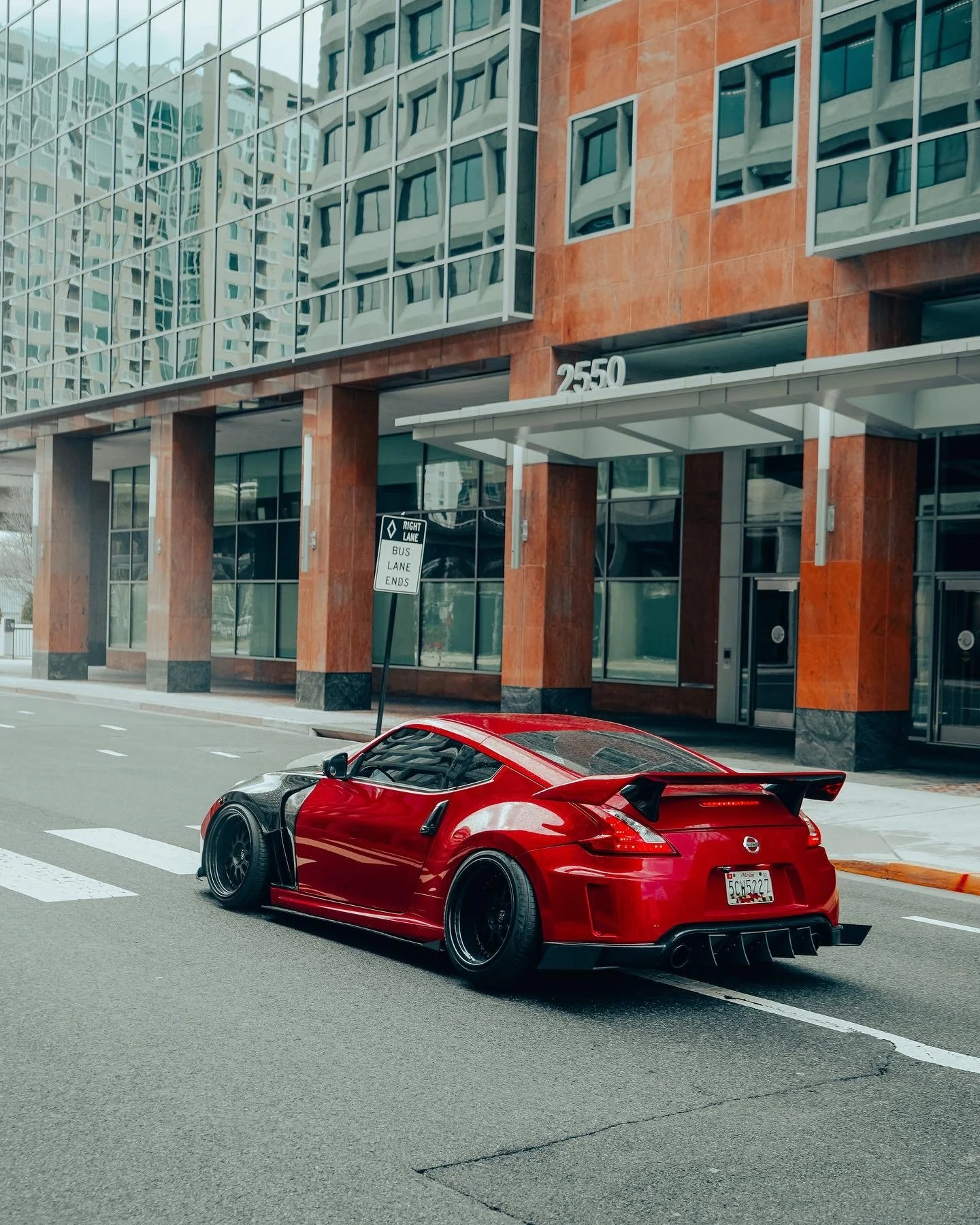 A red sports car with black accents and a large rear wing parked on a city street in front of a modern building with reflective glass windows and stone facade, with a street sign indicating bus lane ends.