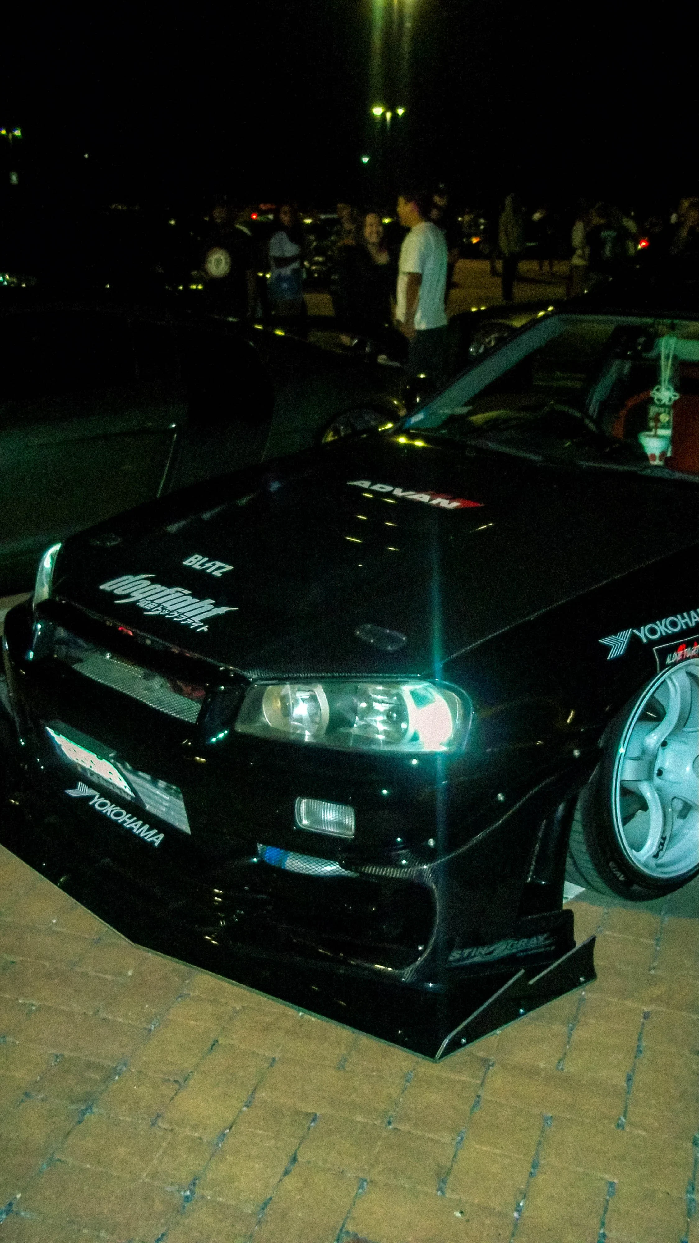 A black race car with Yokohama and Blitz decals, parked at night on a brick surface, with people standing in the background under streetlights.