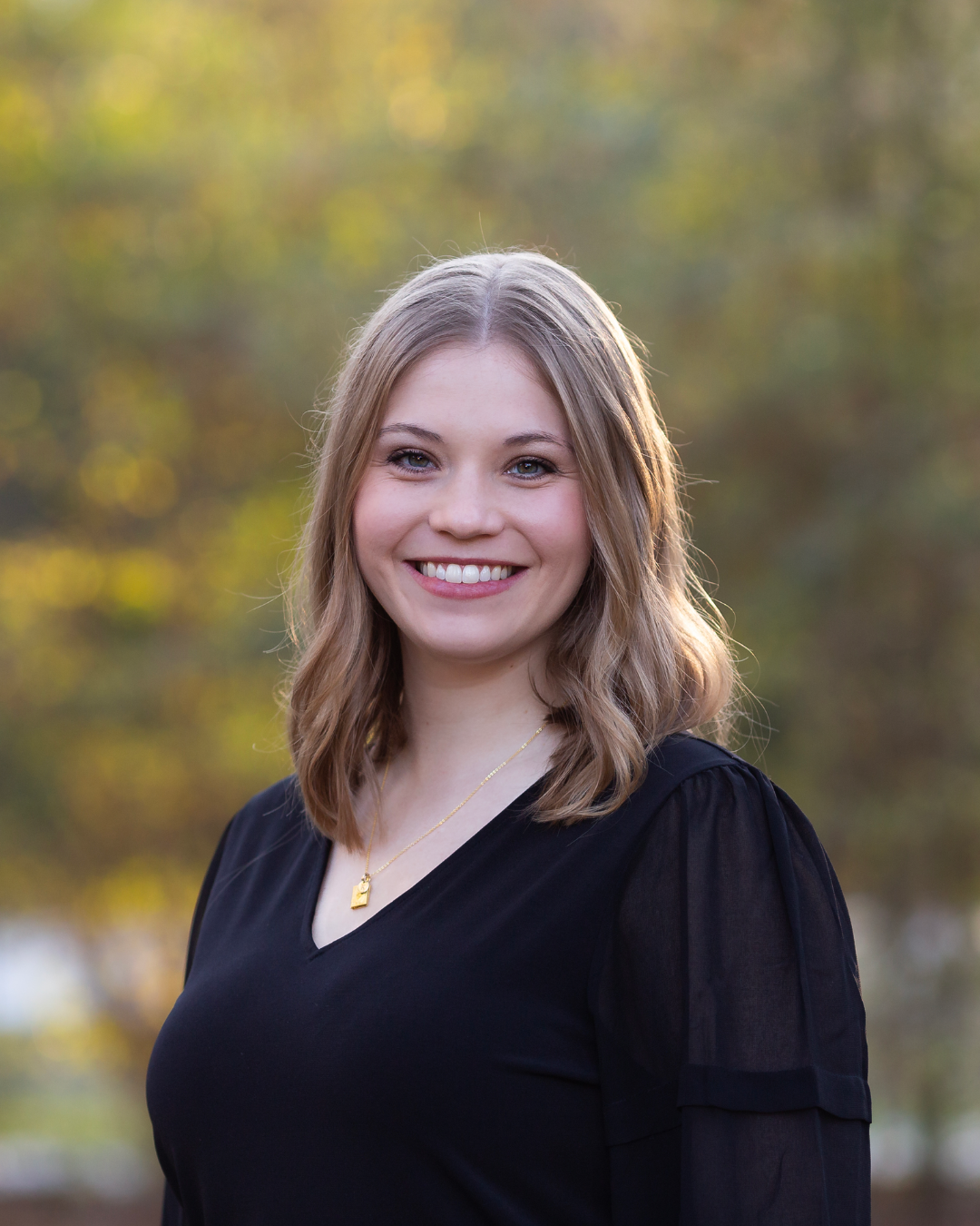 A young woman with shoulder-length light brown hair wearing a black top with puffed sleeves and a gold necklace, smiling outdoors with blurred trees and autumn leaves in the background.