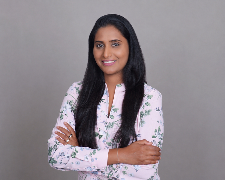 A woman with long black hair wearing a light pink blouse with green leaves, standing against a gray background with arms crossed and smiling.