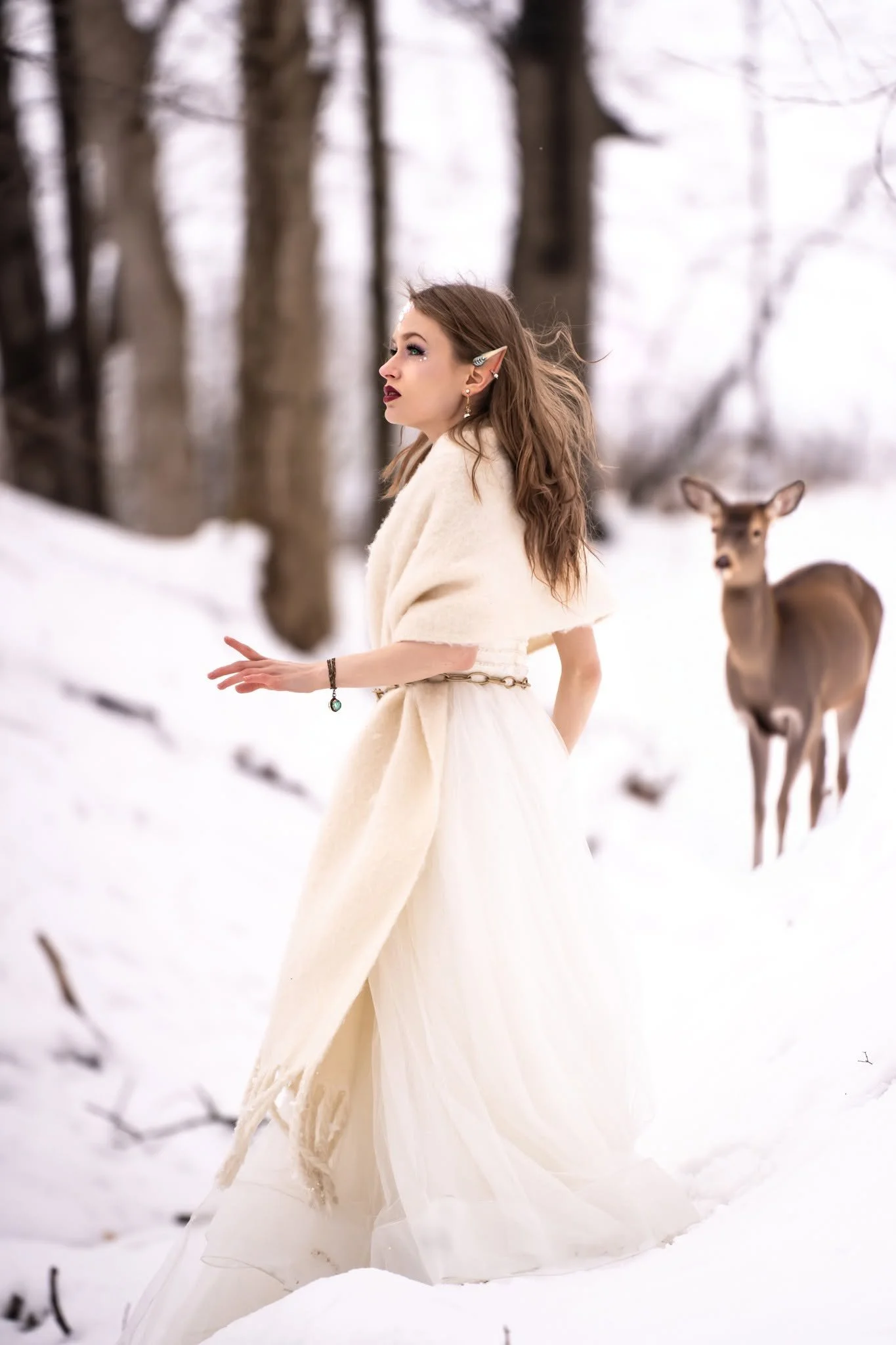 A woman dressed in a flowing white gown with elf-like ears, standing in a snowy forest with a deer nearby.