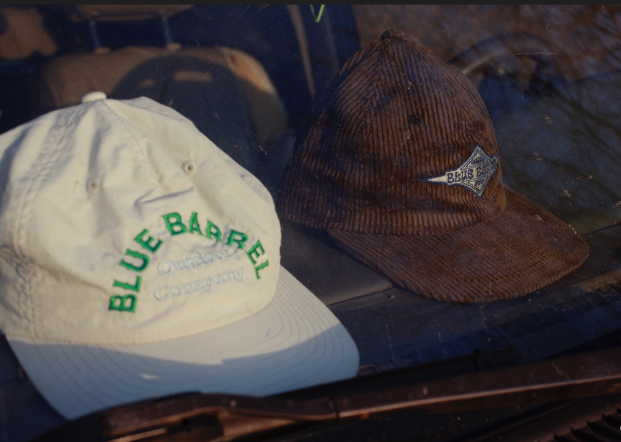Two baseball caps on display behind glass, one white with green stitching reading 'Blue Barrel Outfitter Company,' and one brown with a logo patch.
