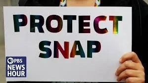 Hand holding a sign that says "PROTECT SNAP" with the PBS NewsHour logo in the bottom left corner.