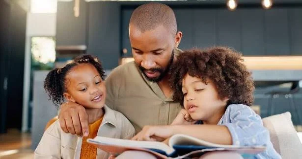 A man reading a book with two young children, one girl and one boy, in a cozy indoor setting.