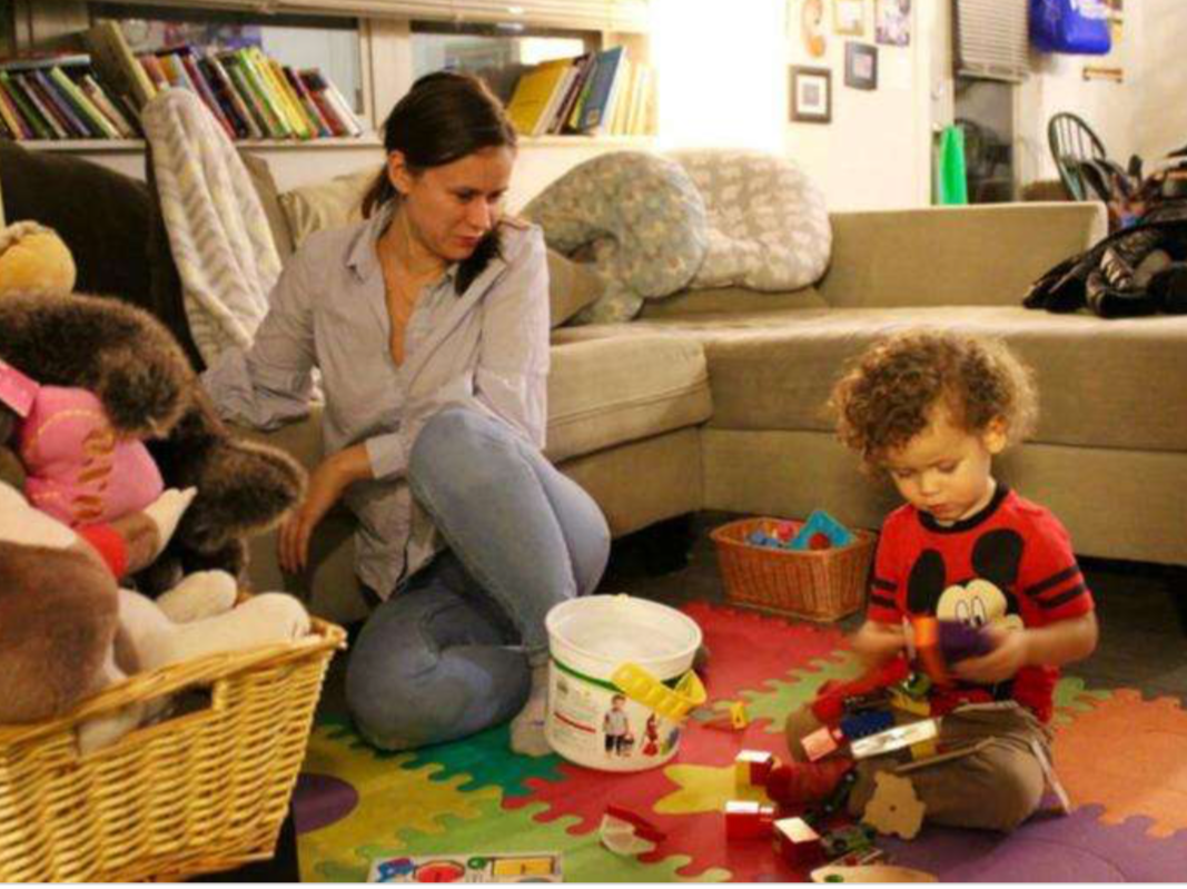 A woman and two children playing with toys on a colorful foam mat in a living room. The woman and one child are sitting on the floor, with the other child near a basket of stuffed animals.