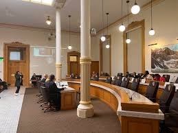 A courtroom with several people seated at a curved bench, microphones, and wooden furnishings, under hanging lights.