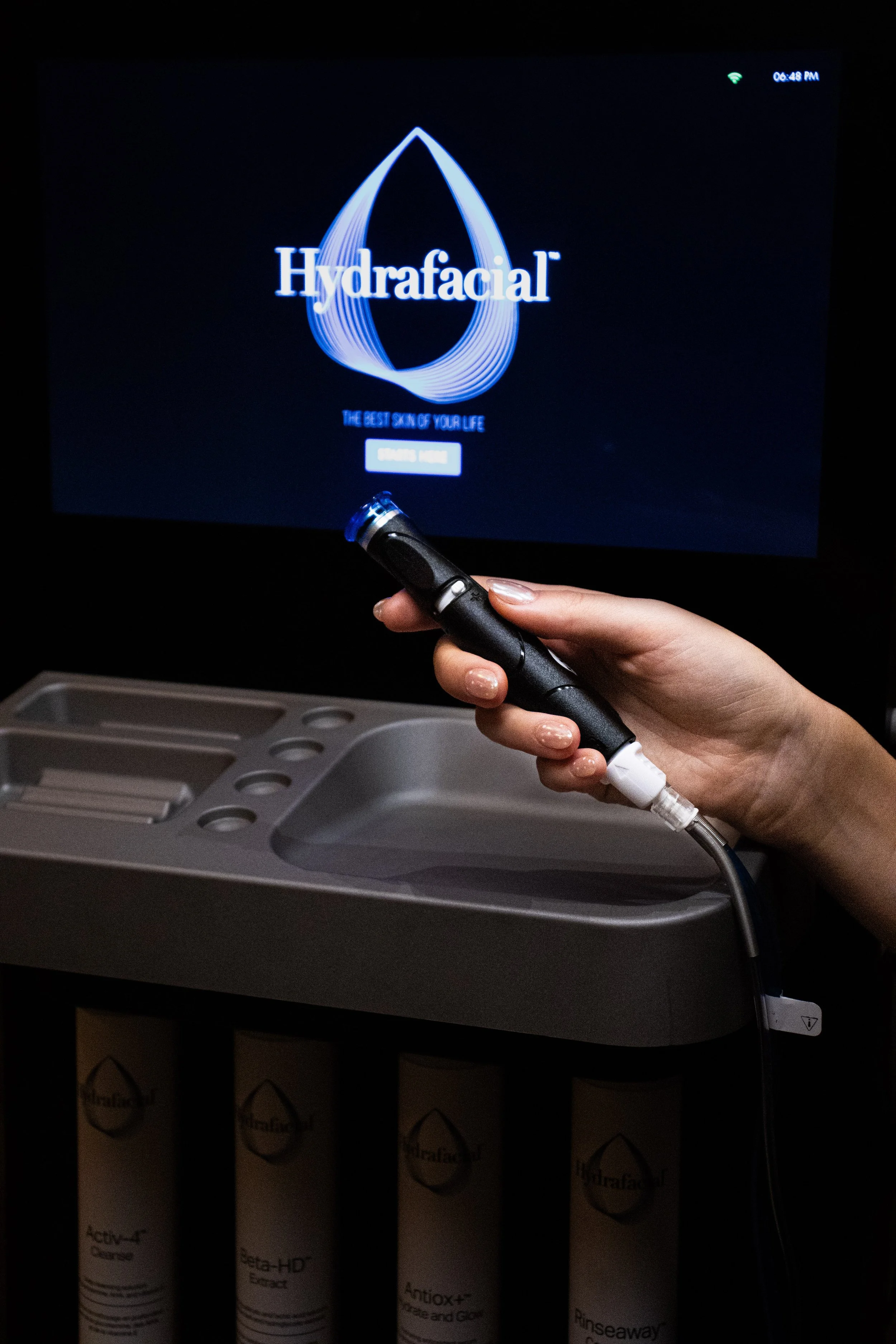 A person holds a black device in front of a computer screen displaying the word 'Hydrafacial' with a water drop graphic and tagline 'The Best Skin of Your Life'; the person is next to a cart with boxed products.