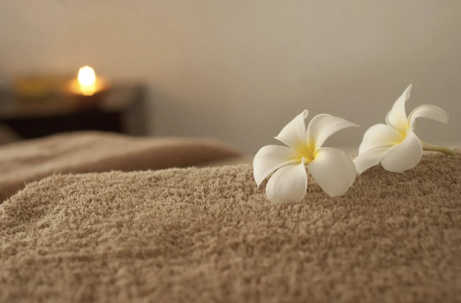 Two white plumeria flowers resting on a soft textured beige surface with a blurred dimly-lit candle in the background.