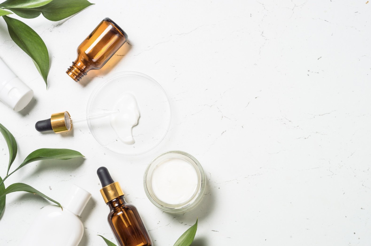 Various skincare products including bottles and droppers, along with green leaves on a white textured surface.