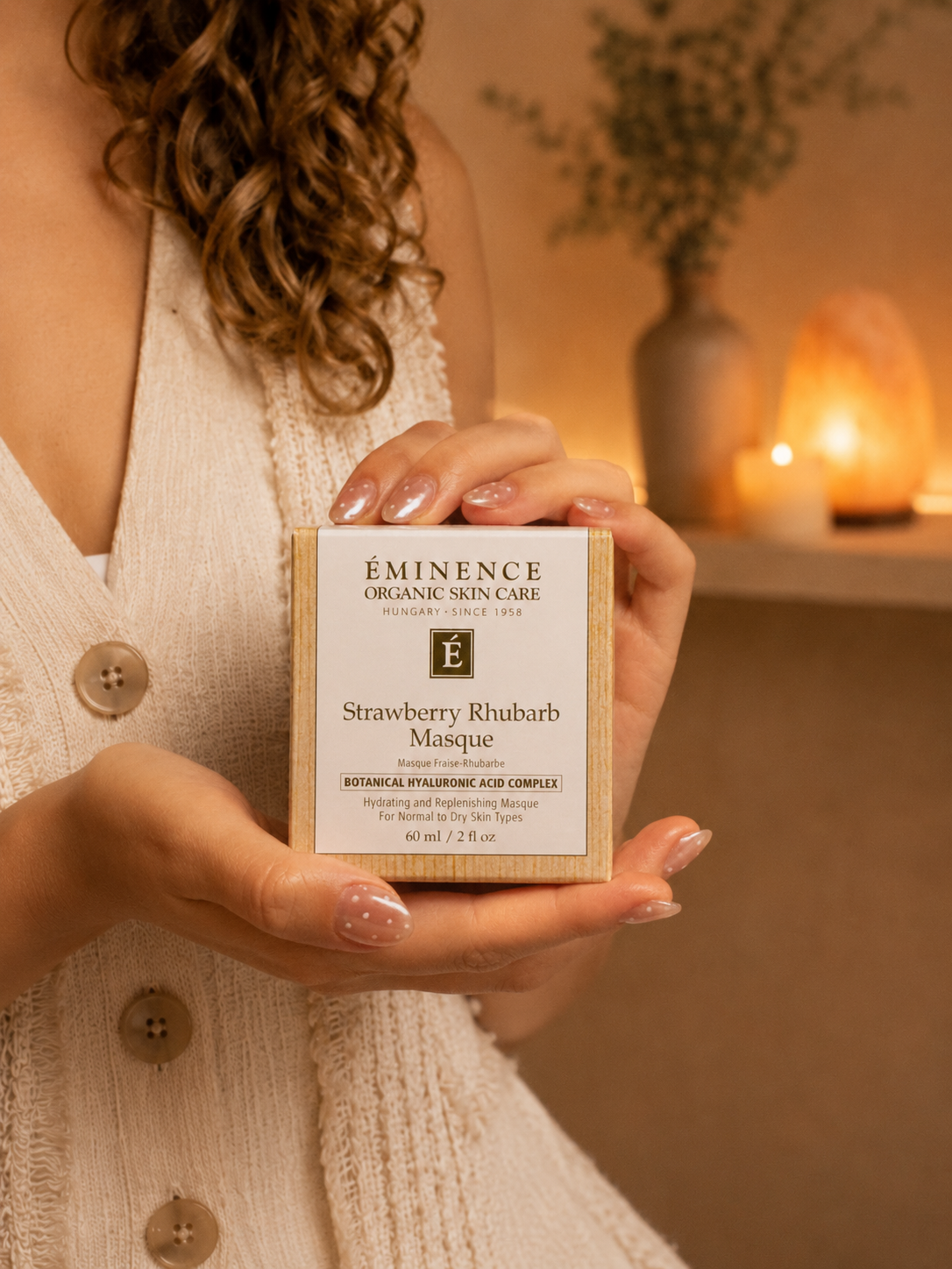 A woman with curly hair holding a box of Eminence Organic Skin Care Strawberry Rhubarb Masque in a cozy, softly lit room, with a vase of flowers and salt lamps in the background.