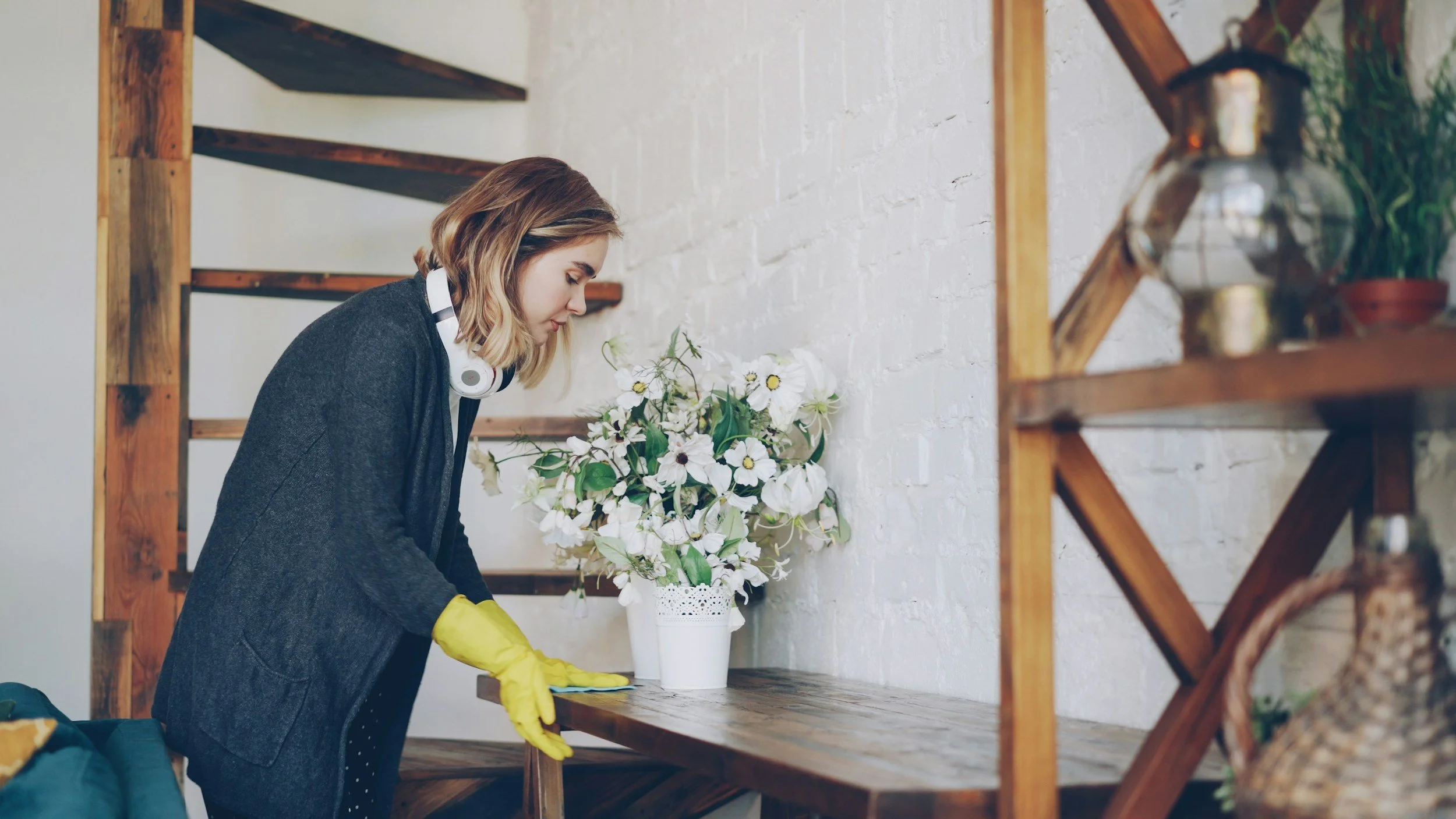 A woman wearing yellow rubber gloves and a dark coat arranging white flowers in a white vase on a wooden table.