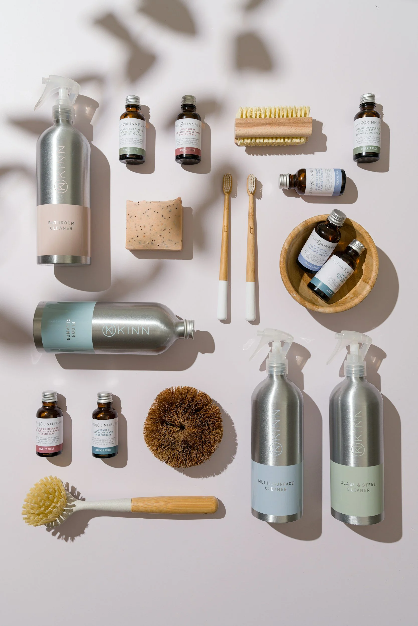 A collection of bathroom cleaning and skincare products, including spray bottles, small bottles, brushes, a soap bar, a loofah, and a cleaning sponge, arranged on a white surface with shadows.