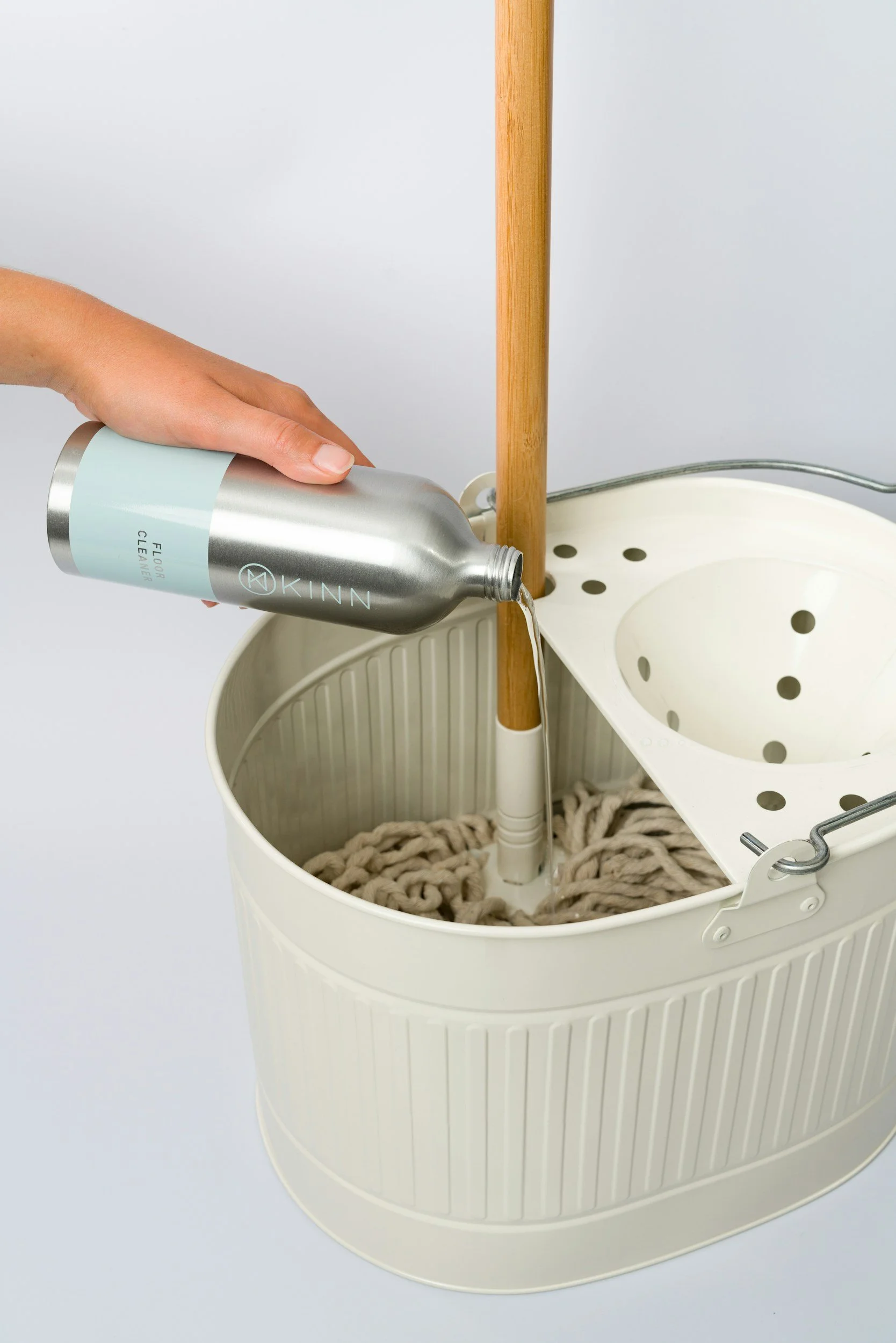 A person pouring floor cleaner into a mop bucket with a built-in wringer, containing a thick mop. The bucket is white with vertical ridges and a wooden mop handle is upright in the wringer.