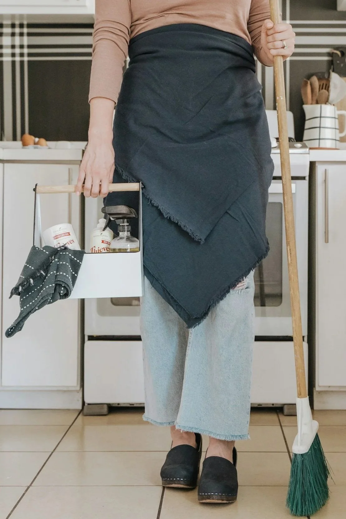 Person standing in a kitchen wearing light-colored jeans, a dark apron, holding a broom, and carrying a cleaning caddy with cleaning supplies and a cloth.