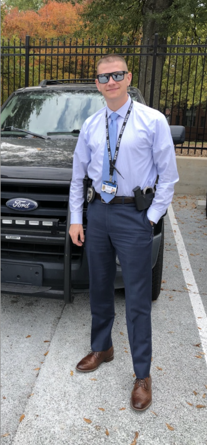 A young man in formal attire, wearing sunglasses and a badge with a lanyard, standing in front of a black Ford vehicle in a parking lot with fall-colored trees in the background.