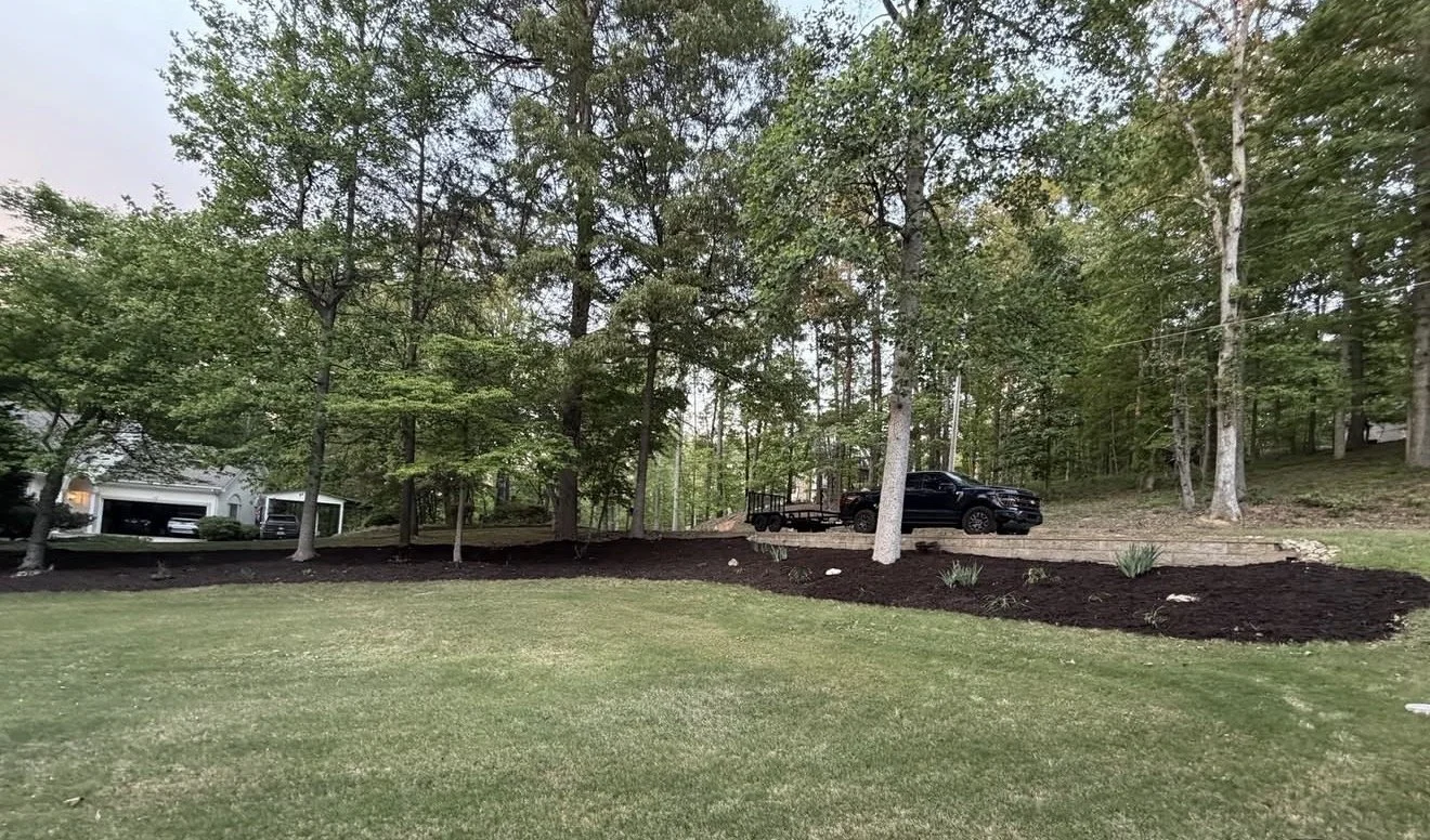 A landscaped backyard with freshly laid dark mulch, green grass, and several trees. There is a black pickup truck with a trailer parked on a dirt pathway among the trees, with a house visible in the background.
