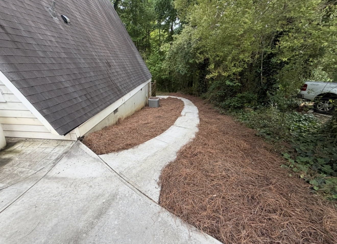 Concrete pathway with pine straw mulch on either side, leading along the side of a house with a sloped shingled roof, surrounded by green trees and bushes, with a parked white truck in the driveway.