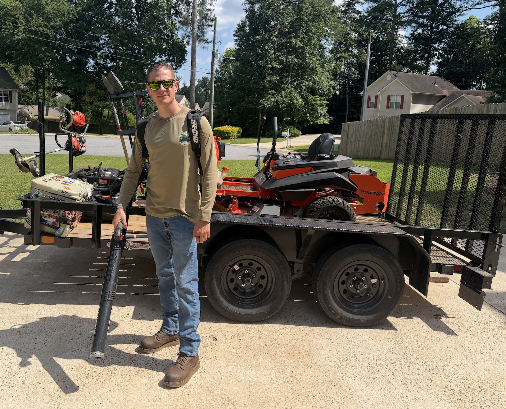 A young man in sunglasses with a backpack holding a leaf blower stands in front of a flatbed trailer carrying a red lawn mower and various tools, parked outdoors in a residential neighborhood with trees, houses, and a wooden fence in the background.