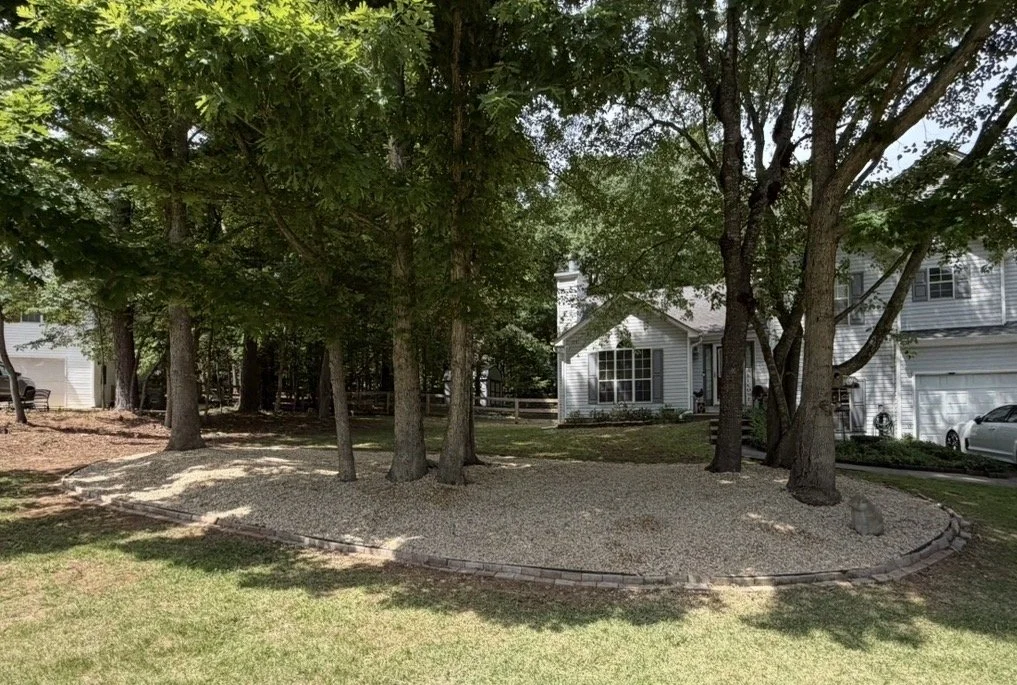 Residential backyard yard with trees, stone border, and gravel ground, with a house and a car in the background.