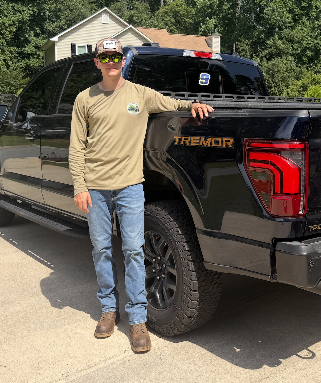 A man wearing sunglasses, a beige long-sleeve shirt, blue jeans, and brown boots standing next to a black Ford F-150 Tremor pickup truck, resting his arm on the truck, in a suburban driveway with a beige house and trees in the background.