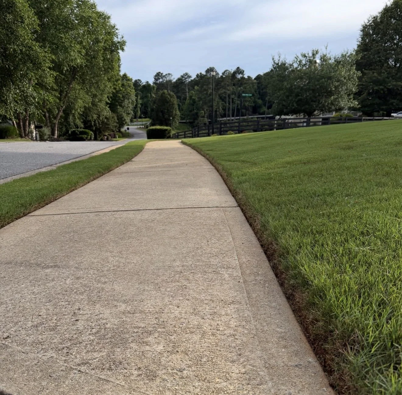 A sidewalk bordered by grass on one side and a street with trees on the other in a suburban neighborhood.