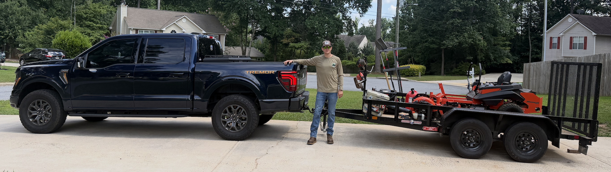 A man stands next to a black pickup truck with a trailer attached, carrying a small orange and black lawnmower. The scene takes place on a residential street with houses and trees in the background.