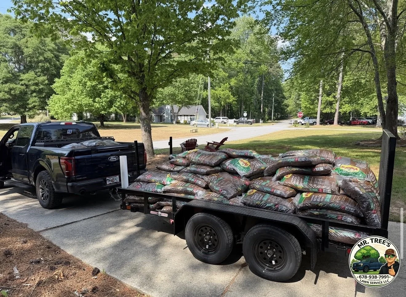 A black pickup truck attached to a flatbed trailer loaded with numerous bags of mulch, parked on a driveway in a residential neighborhood with green trees and houses in the background.