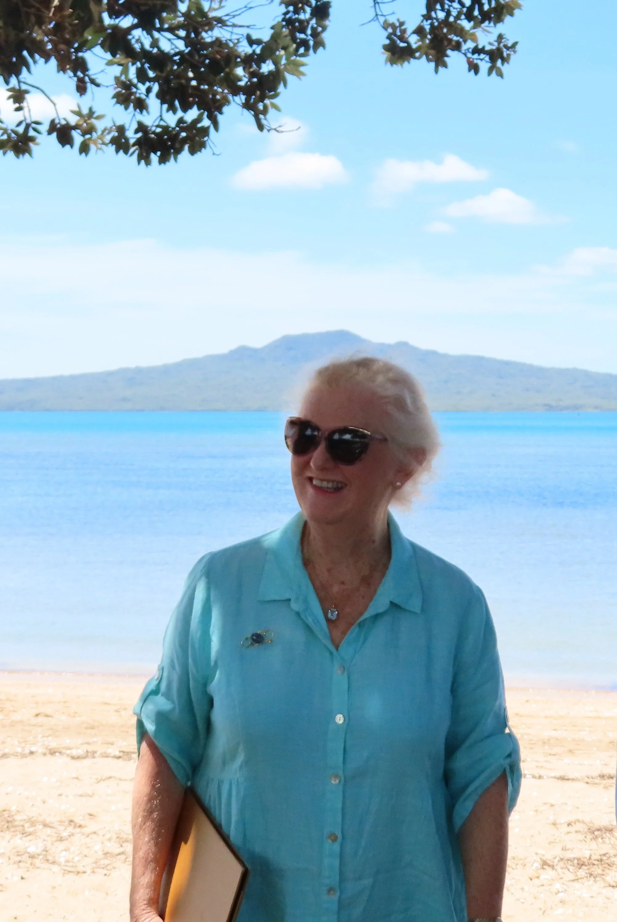 Barbara Johnson celebrant, wearing sunglasses and a light blue button-up shirt, smiling at the camera, standing on a beach with ocean, mountains, and blue sky in the background.