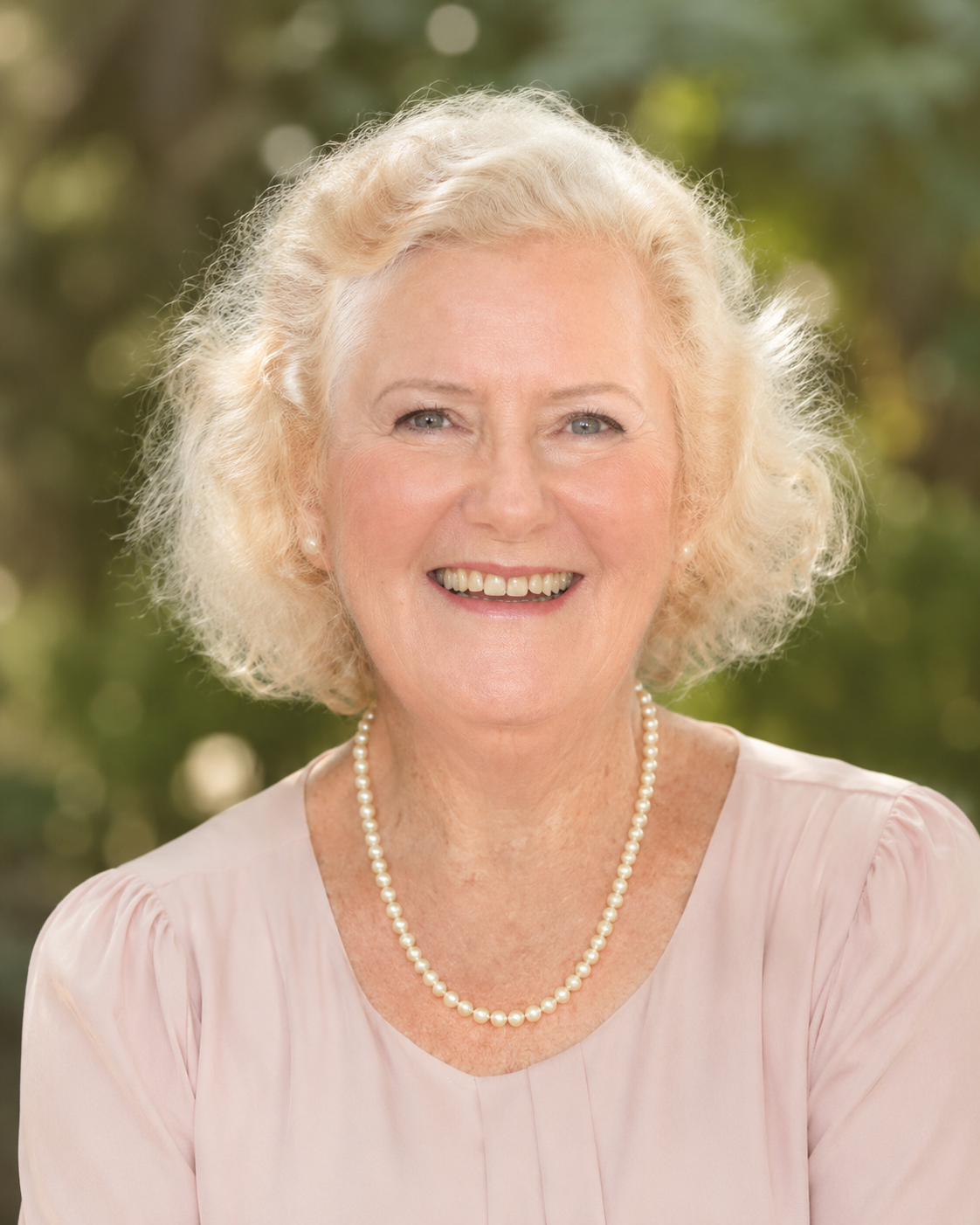 A portrait of an elderly woman with curly blonde hair, wearing a light pink blouse, a pearl necklace, and pearl earrings, smiling outdoors with blurred green trees in the background.