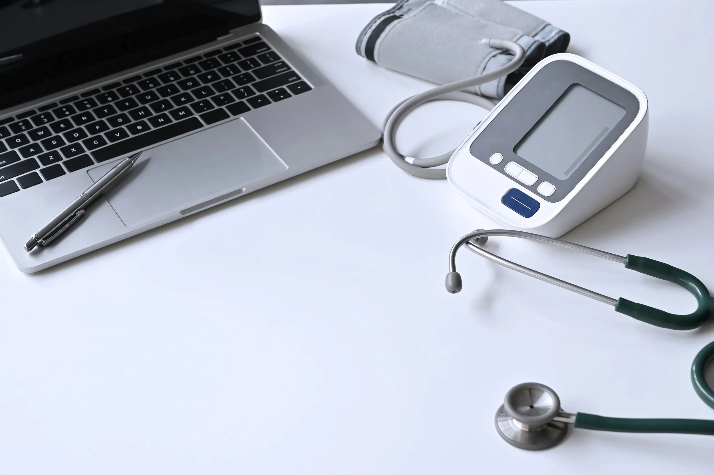 A desk with a Mac-style laptop, a pen, a blood pressure monitor, and a stethoscope on a white surface.