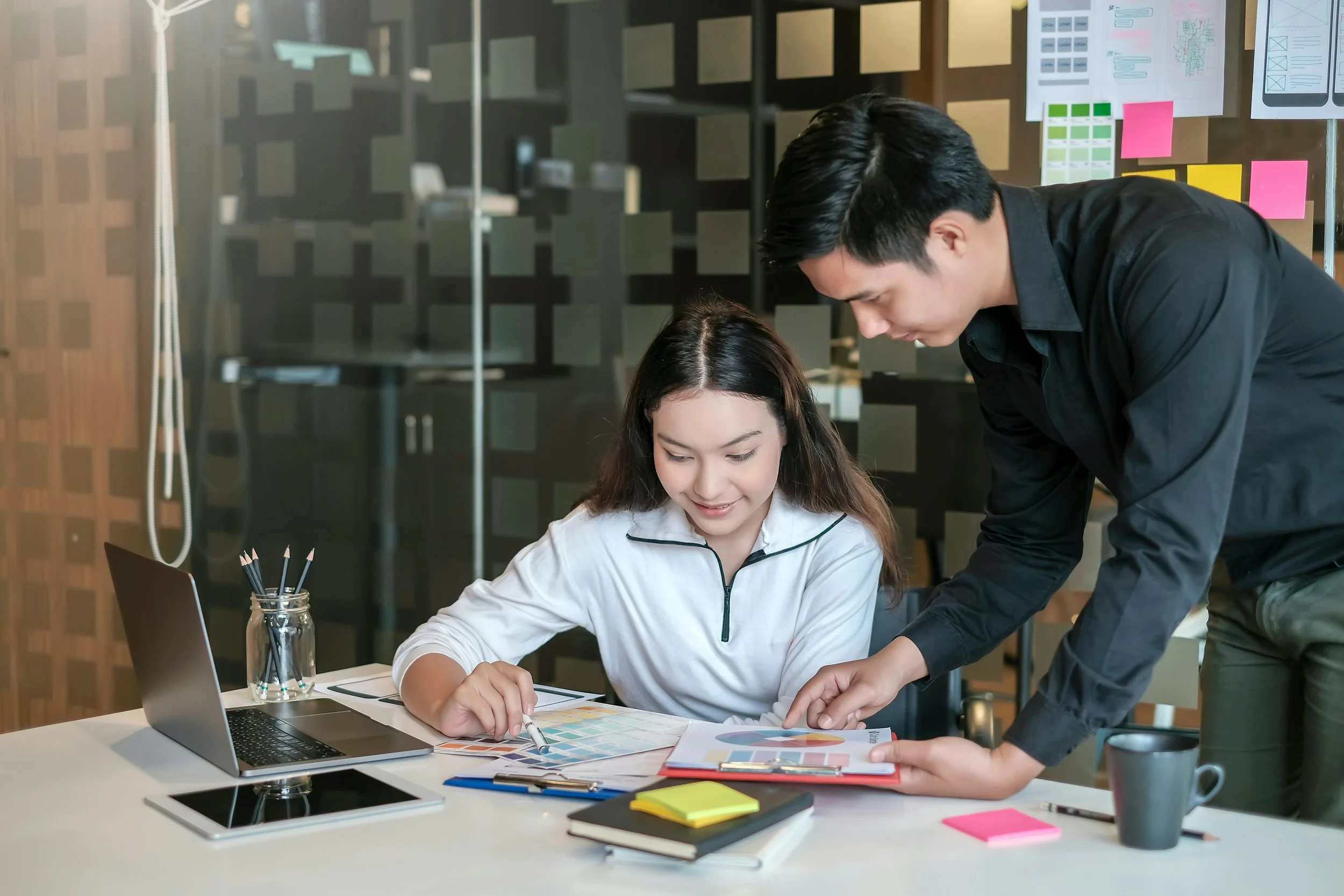 Two app designers, a sitting female and standing male, working collaboratively at a table, with the male looking down at the user interface diagrams shown to him by the female.  The desk has a variety of pastel Post-It Notes along with hand drawings.