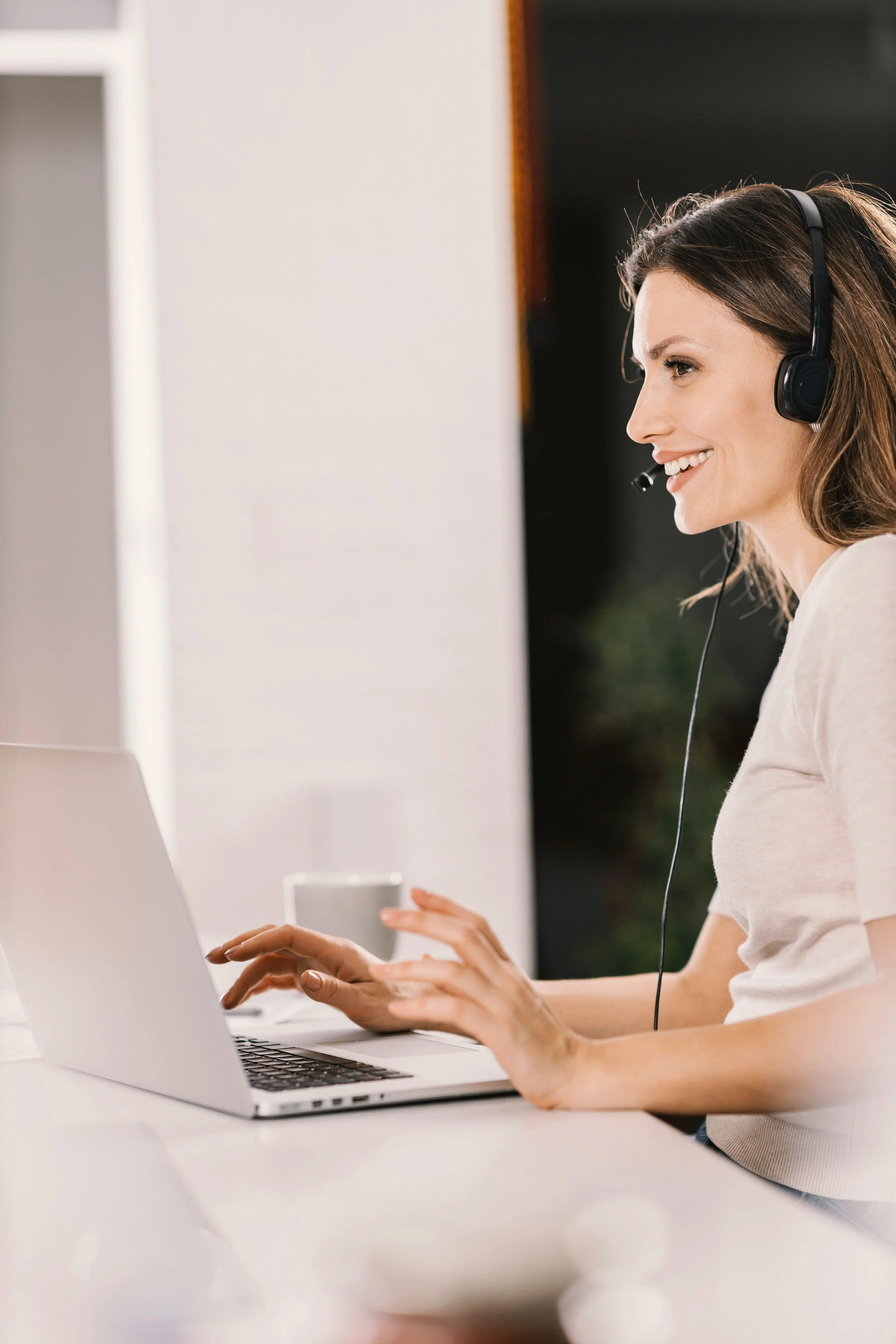 A woman smiling while working on a laptop, wearing a headset with a microphone, sitting at a white desk in a bright room.