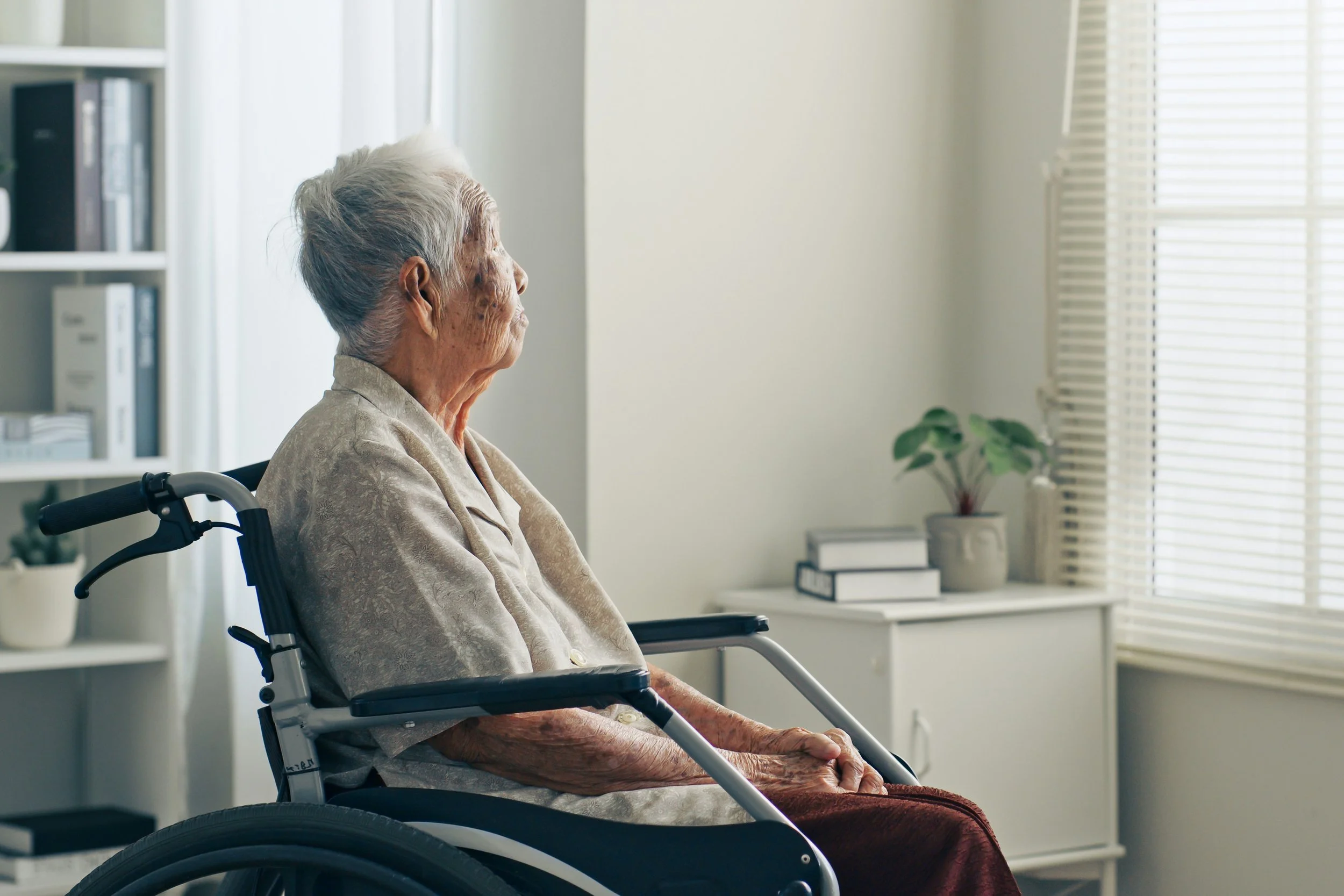 An elderly woman in a wheelchair, sitting and looking out the window in a bright, living room with blinds, a small desk, and a potted plant.