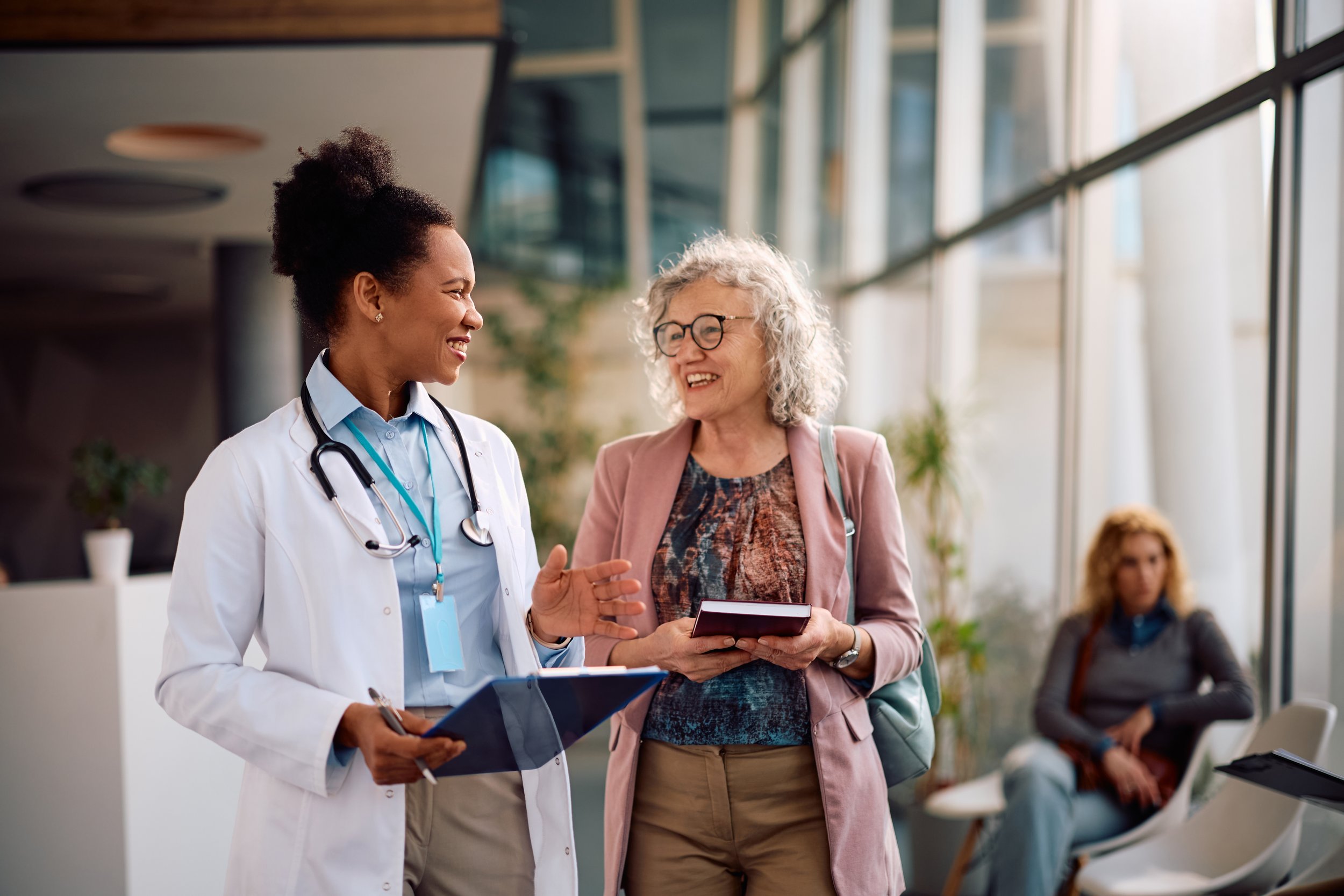 A female doctor with a stethoscope and clipboard talking to an older woman in a pink blazer holding a tablet, both smiling, in a modern indoor setting with other seated people in the background.
