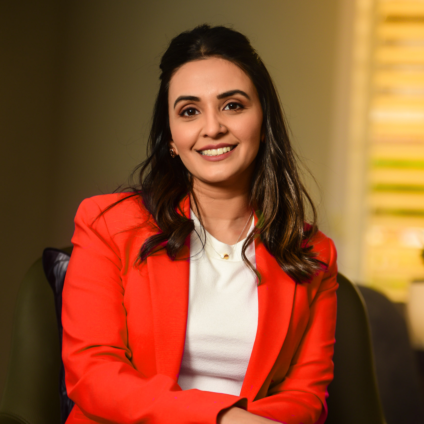 A smiling woman with long dark hair, wearing a red blazer and a white top, sitting in a chair with a blurred background and horizontal blinds on the right side.