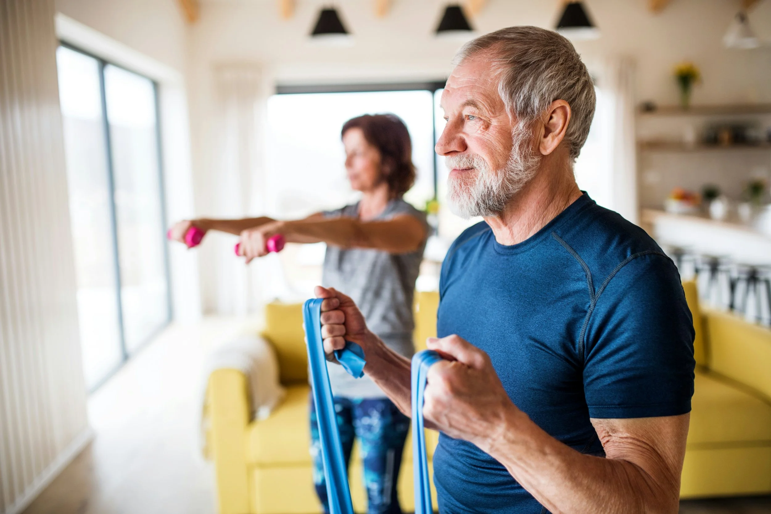 An elderly man and woman exercising indoors, the man holding a resistance band and the woman stretching with pink dumbbells in her hands. Bright, modern room with large windows and yellow furniture.