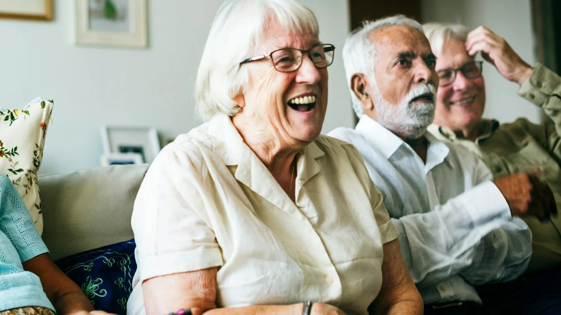 Four elderly people sitting on a couch, smiling and laughing, in a living room setting.