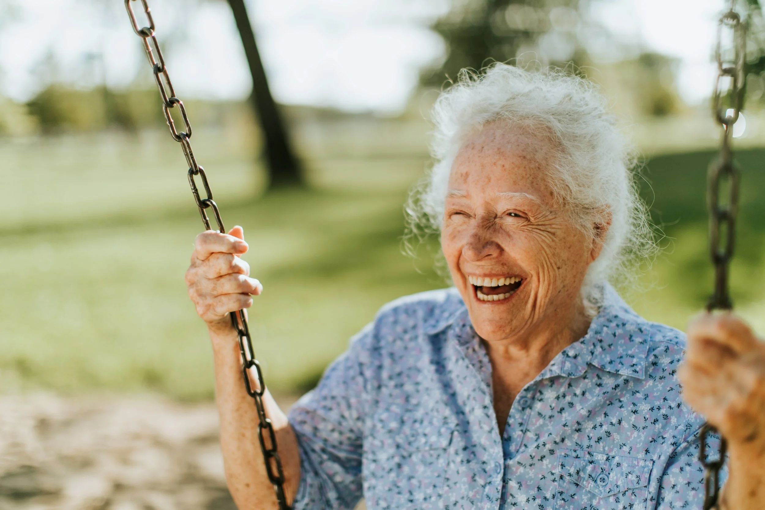 An elderly woman with white curly hair smiling while sitting on a swing outdoors, holding the chains with both hands, in a park with green trees and grass in the background.